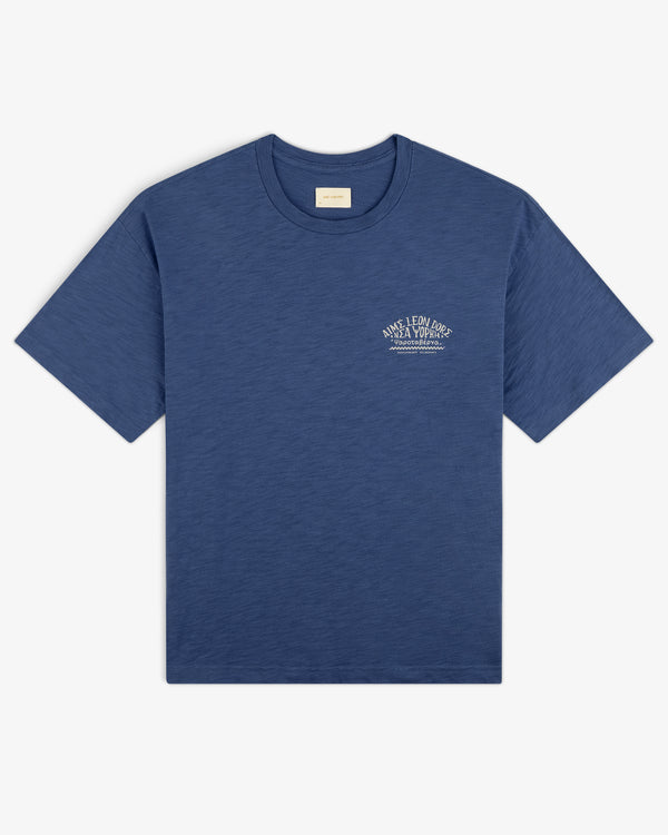 Taverna Tee: Blue short-sleeve t-shirt with small white text graphic on left chest, shown on white background.