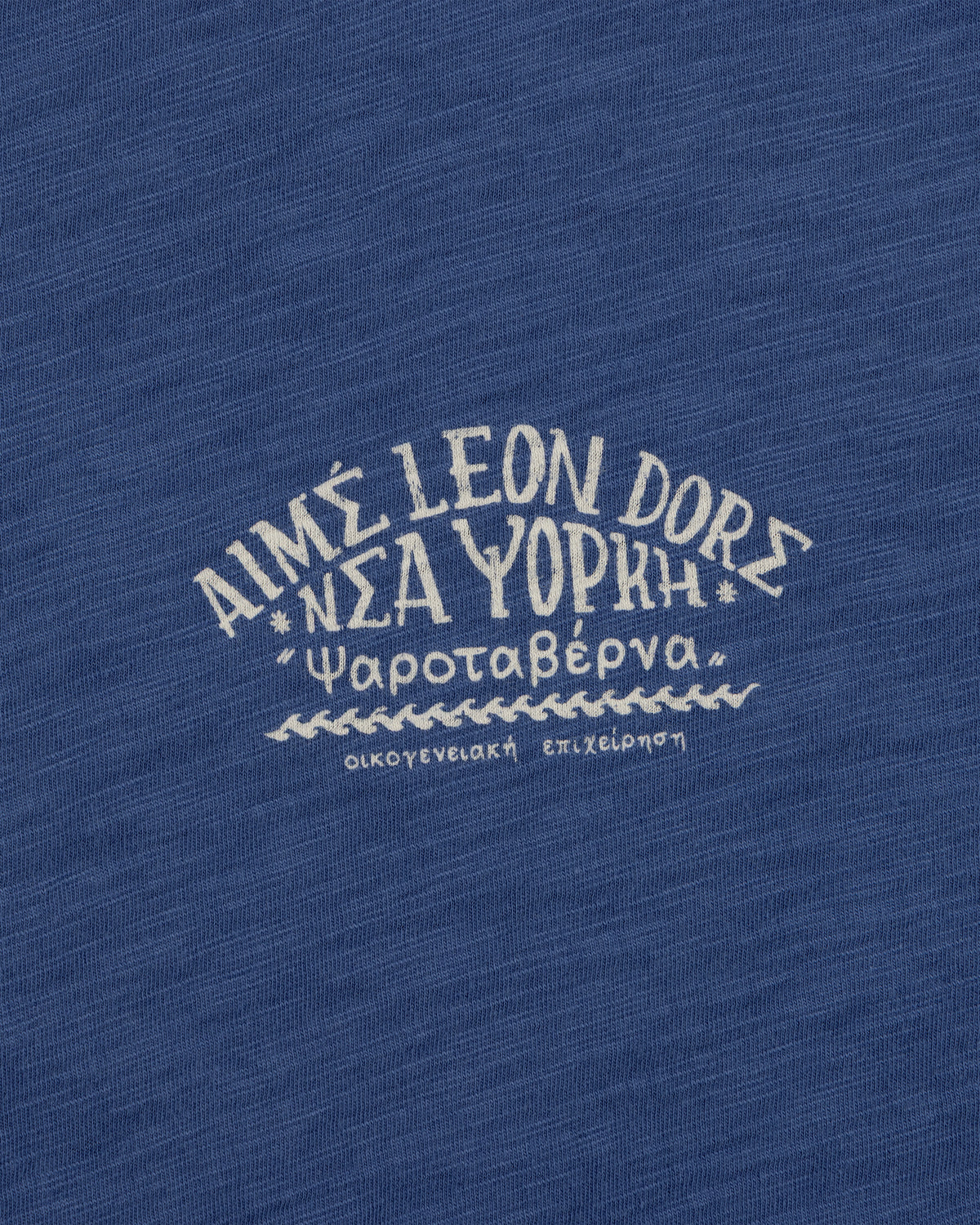 White Greek text and AIMÉ LEON DORE on blue textured fabric, featured on the Taverna Tee.