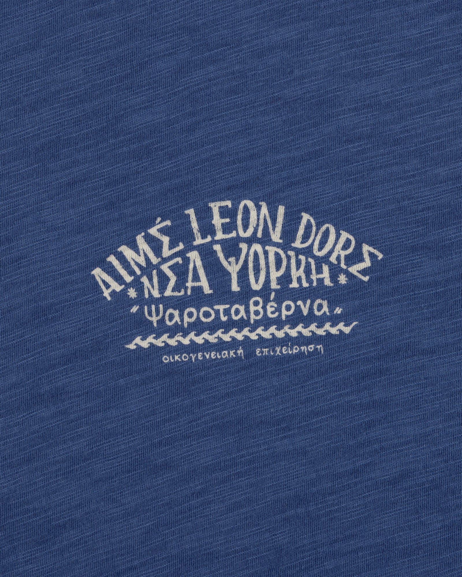 White Greek text and AIMÉ LEON DORE on blue textured fabric, featured on the Taverna Tee.