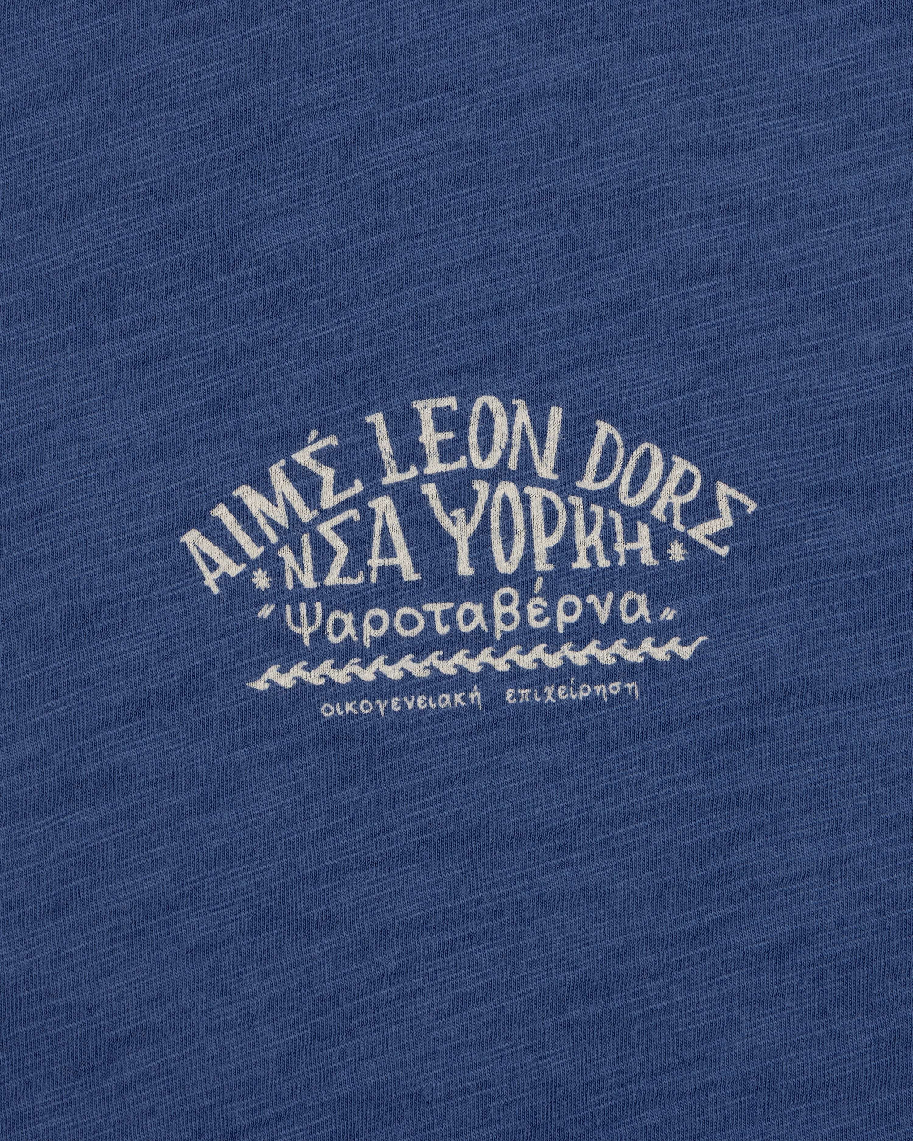 White Greek text and AIMÉ LEON DORE on blue textured fabric, featured on the Taverna Tee.