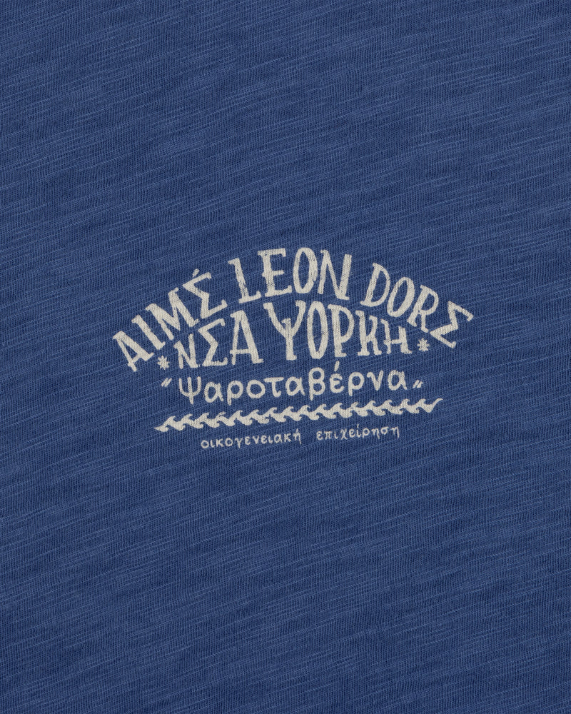 White Greek text and AIMÉ LEON DORE on blue textured fabric, featured on the Taverna Tee.