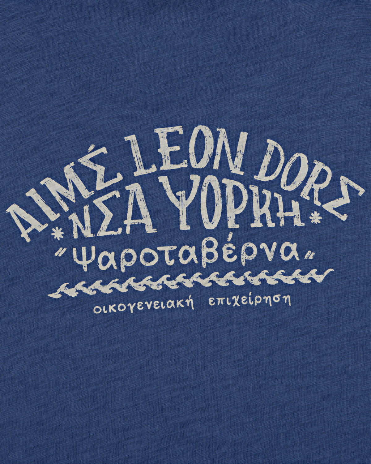 The Taverna Tee features blue fabric with white Greek and English text in a handwritten style.