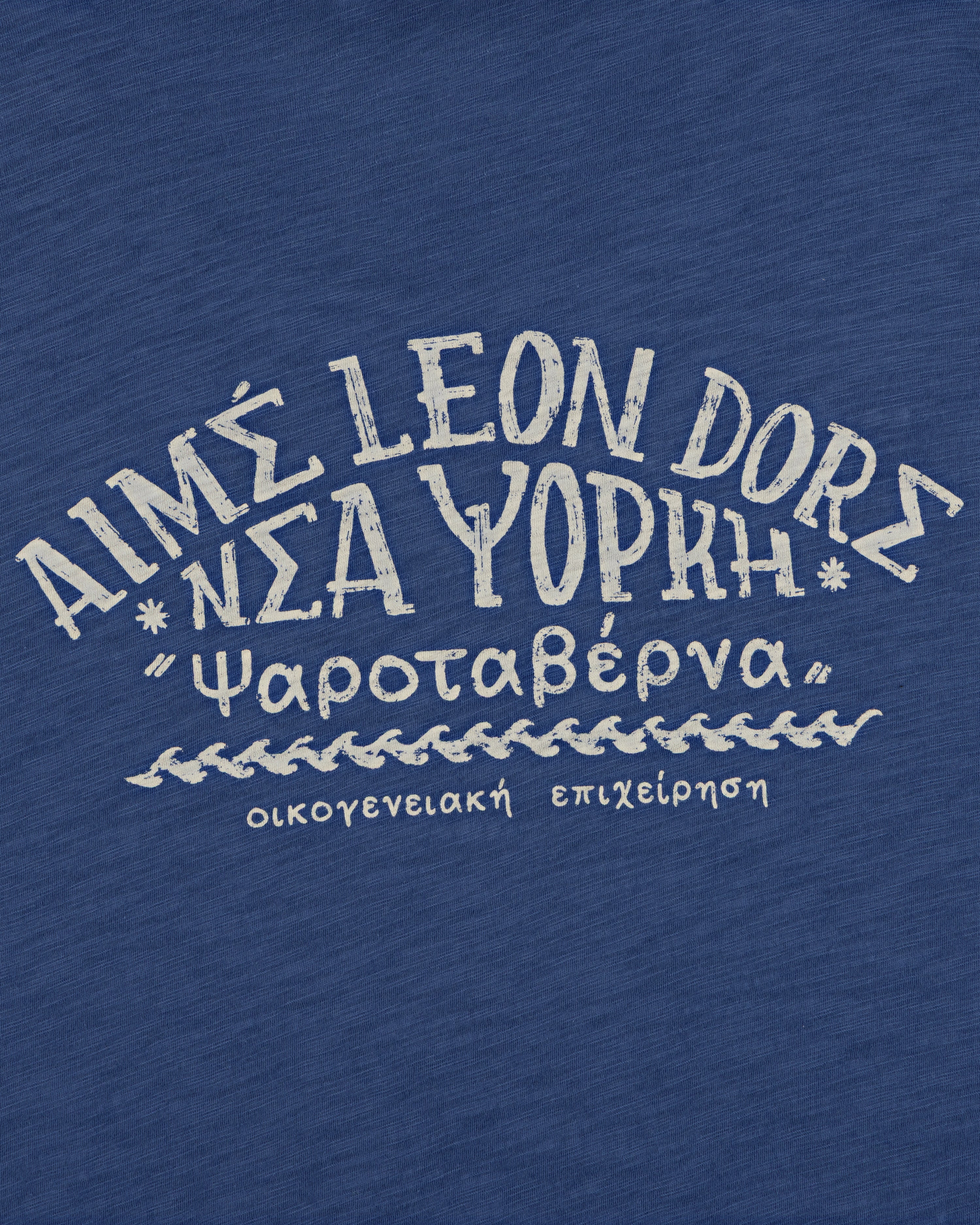 The Taverna Tee features blue fabric with white Greek and English text in a handwritten style.