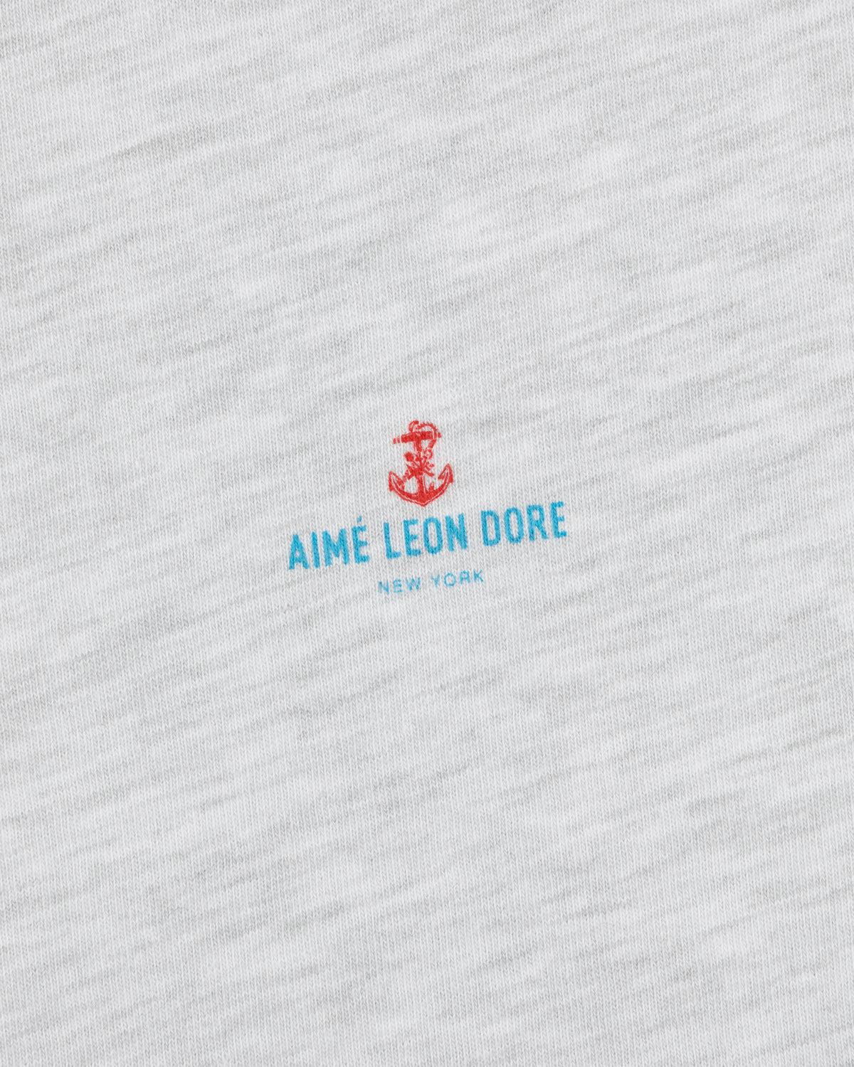 Close-up of the Coastal Flag Tee's white fabric with a small red anchor and blue AIMÉ LEON DORE NEW YORK text.