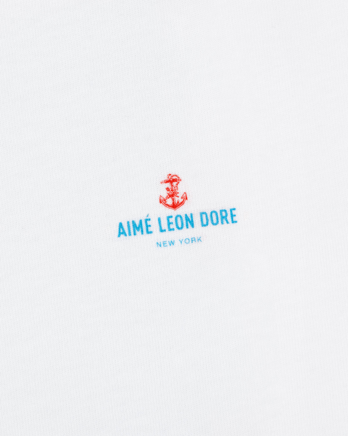 White fabric featuring a small red anchor above blue "AIMÉ LEON DORE NEW YORK" text. Coastal Flag Tee.
