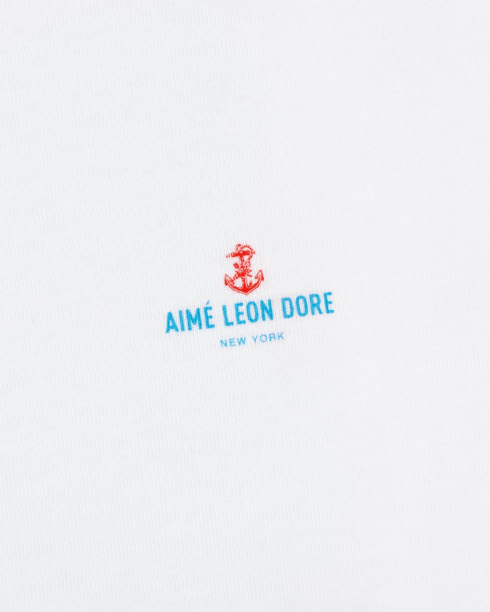 White fabric featuring a small red anchor above blue "AIMÉ LEON DORE NEW YORK" text. Coastal Flag Tee.