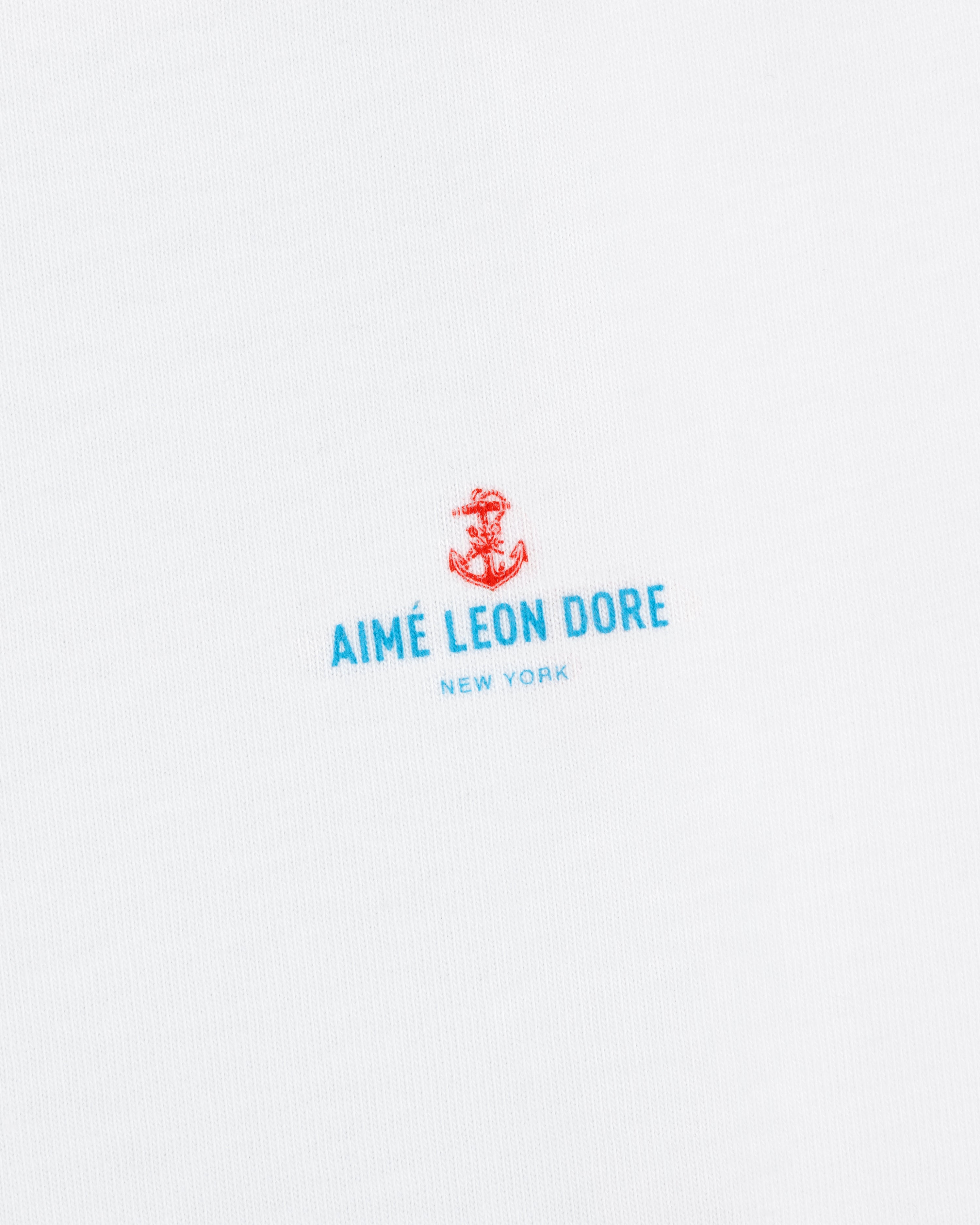 White fabric featuring a small red anchor above blue "AIMÉ LEON DORE NEW YORK" text. Coastal Flag Tee.