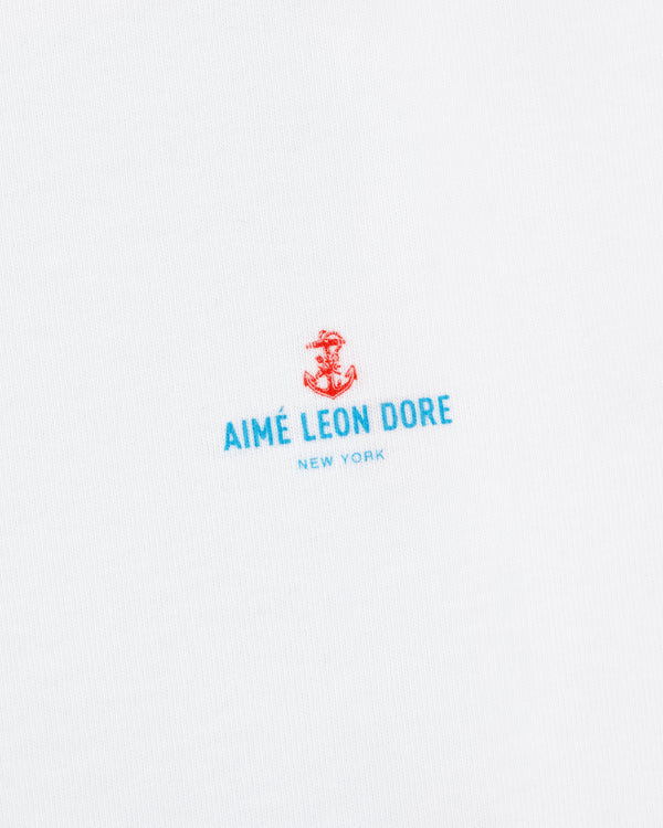 White fabric featuring a small red anchor above blue "AIMÉ LEON DORE NEW YORK" text. Coastal Flag Tee.