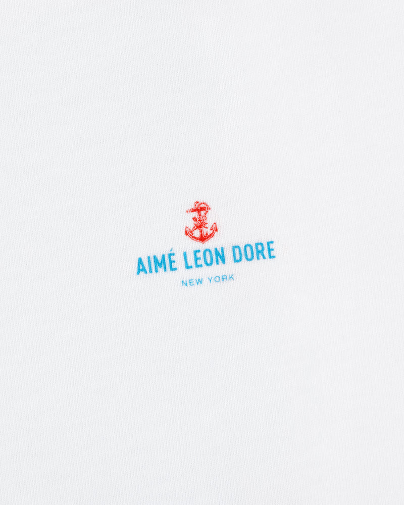 White fabric featuring a small red anchor above blue "AIMÉ LEON DORE NEW YORK" text. Coastal Flag Tee.