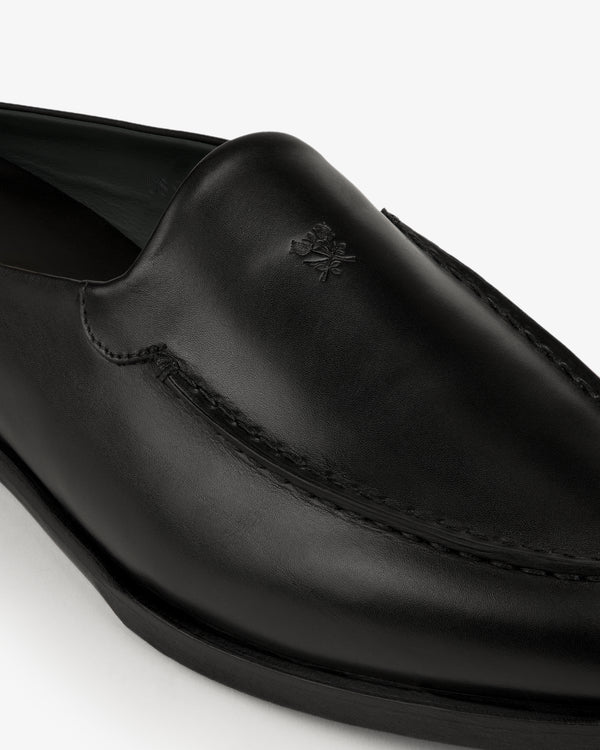 Close-up of the Venetian Mule, a black leather loafer with visible stitching and a small embossed logo on the upper.