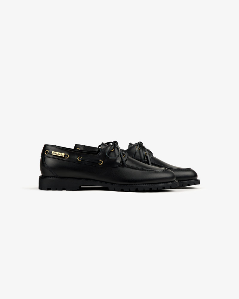 Pápia Boat Shoe in black leather with laces and gold eyelets, side view, on a white background.