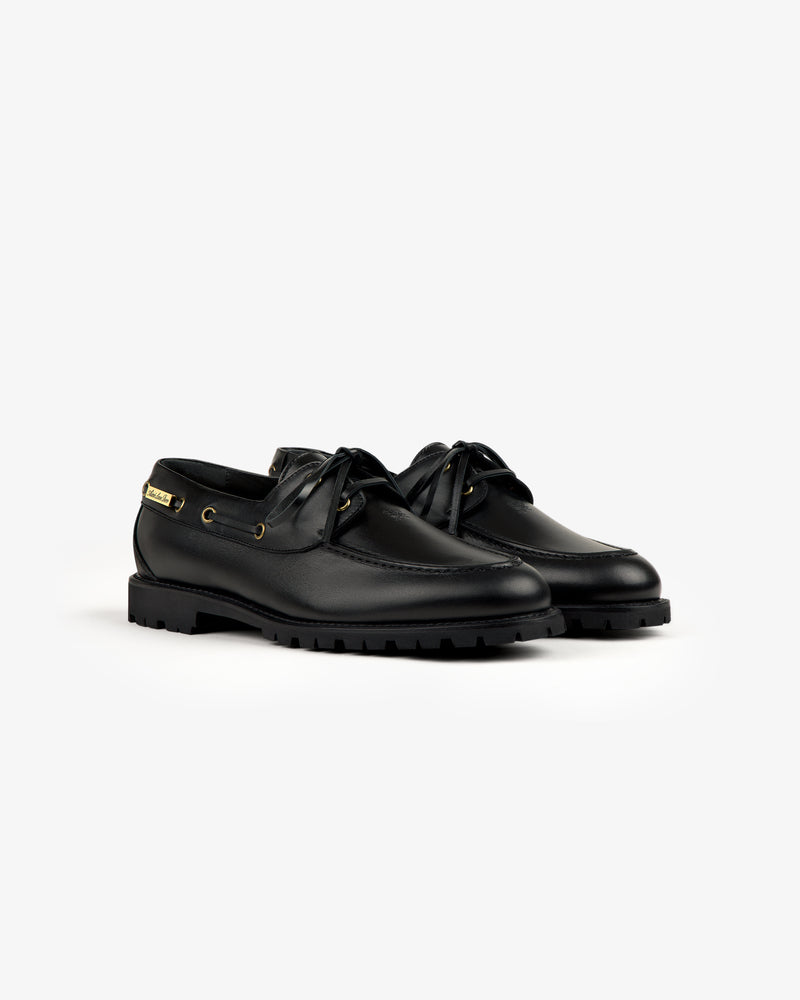 Pápia Boat Shoe: black leather boat shoes with laces and gold eyelets, shown on a white background.