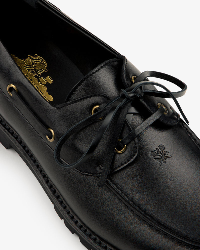Close-up of the Pápia Boat Shoe: black leather loafer with laces, gold eyelets, and embossed logo by the toe.