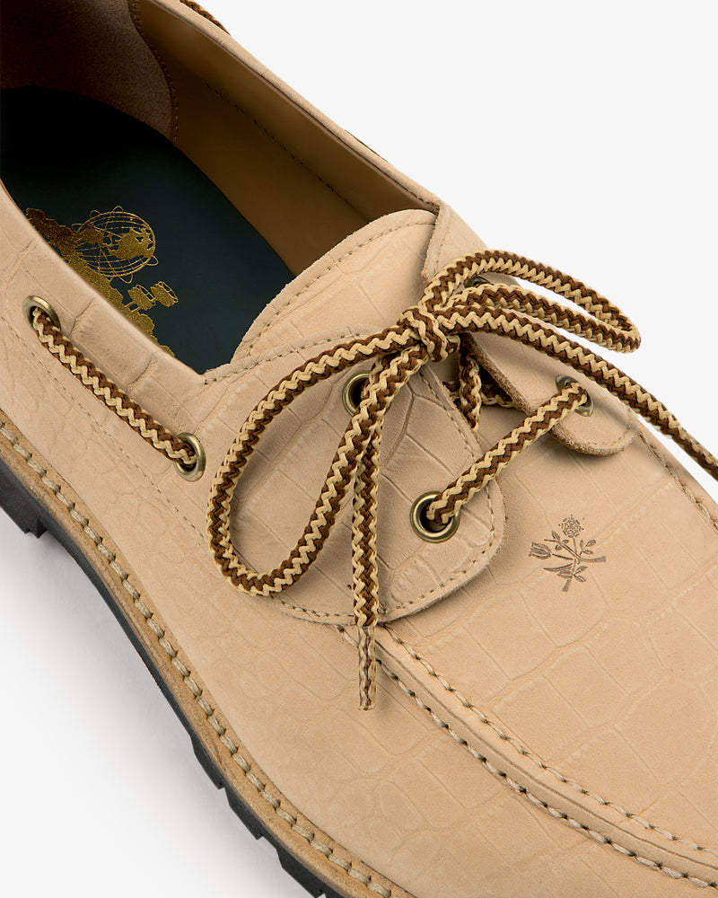 The Pápia Boat Shoe is beige leather with a textured pattern, braided laces, and an embossed emblem near the laces.