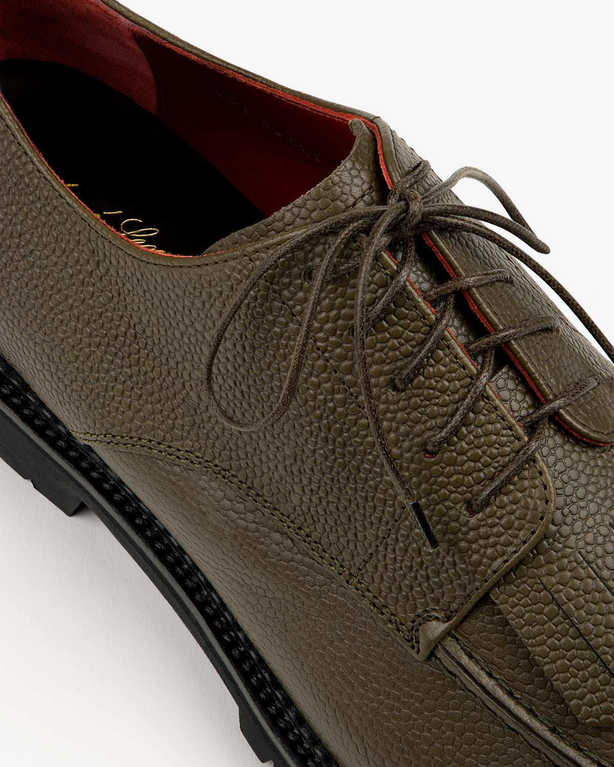 Close-up of a textured olive green Leather Lug Sole Derby with black sole and red interior lining.