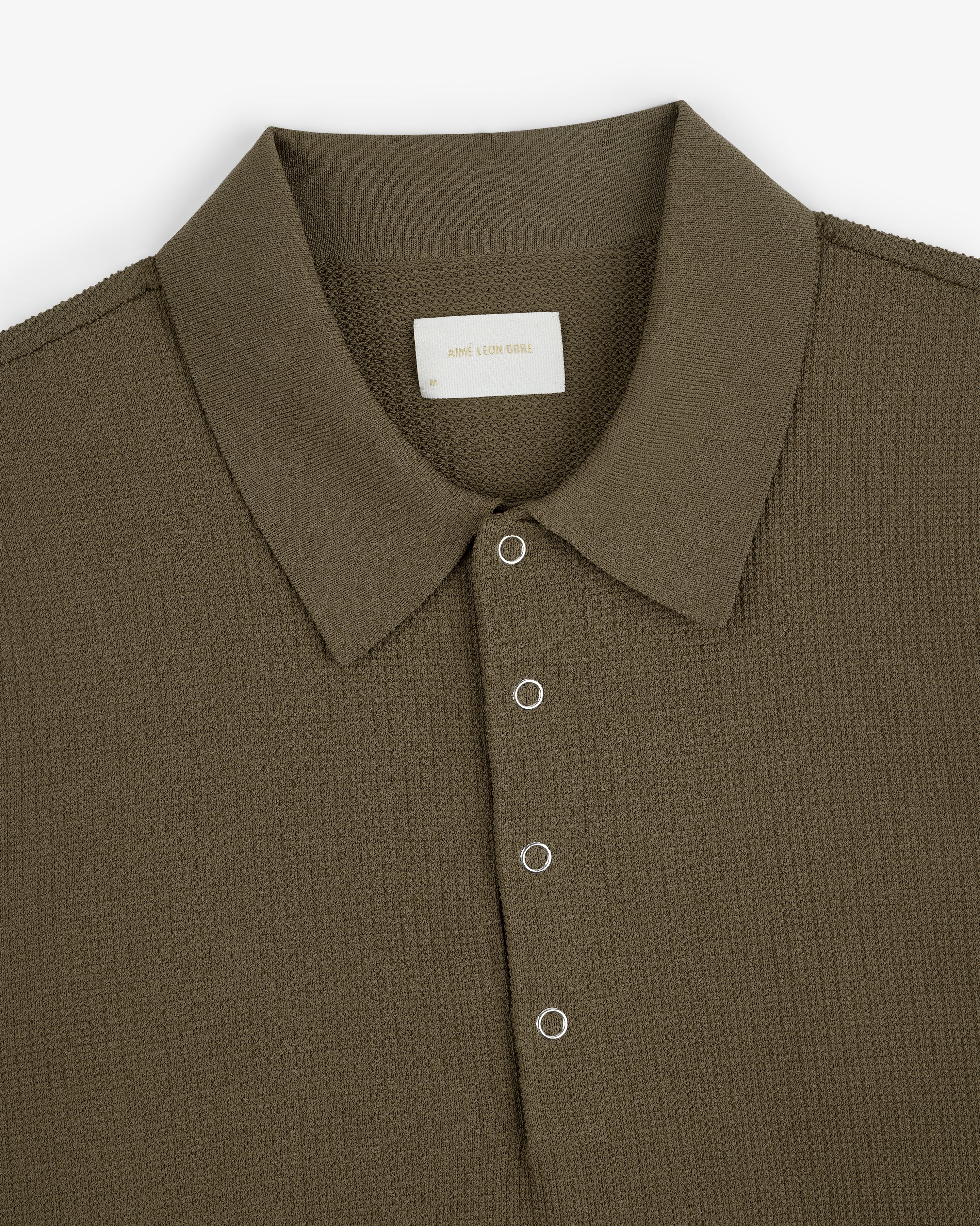 Olive green Knit Polo with textured fabric, spread collar, and four silver eyelet buttons on the placket.