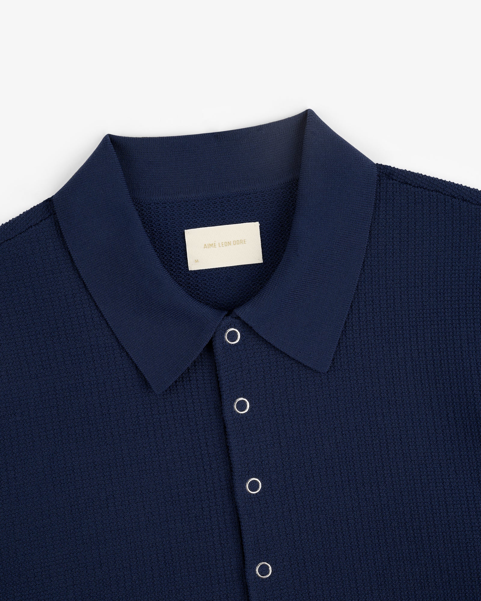 Navy blue Knit Polo with silver snap buttons and a visible Aimé Leon Dore label on the inside collar.