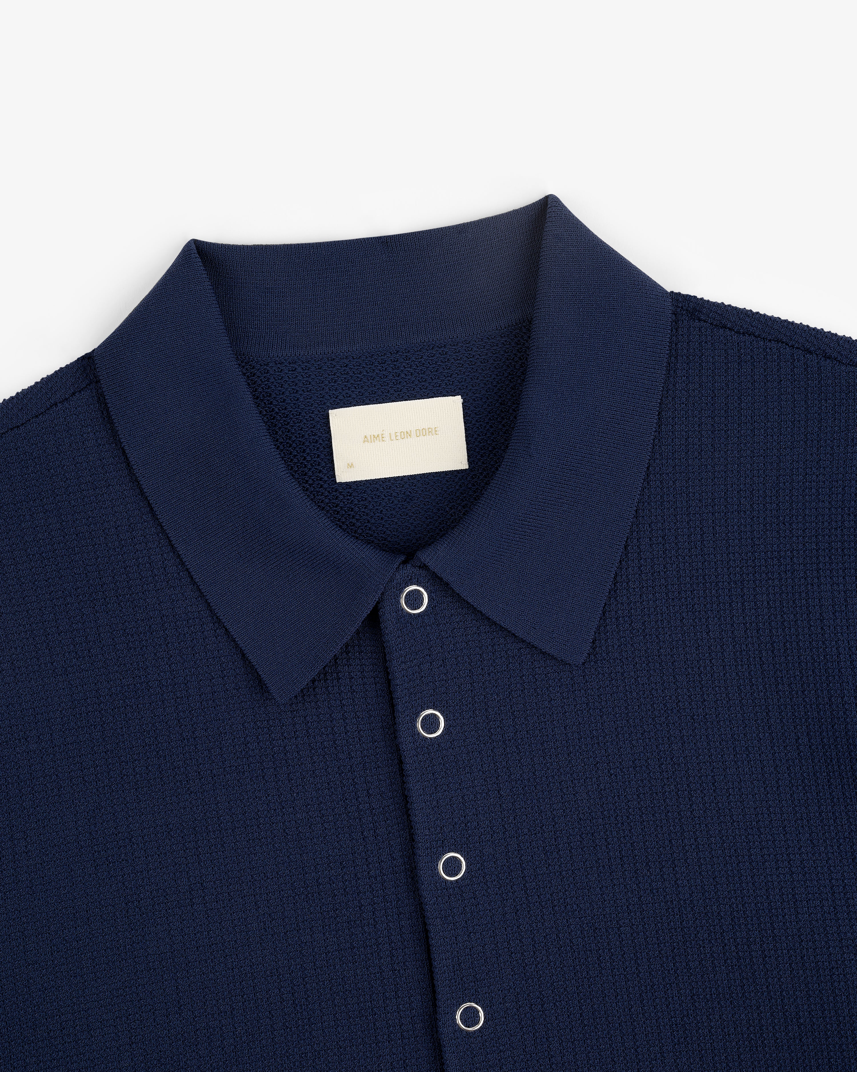 Navy blue Knit Polo with silver snap buttons and a visible Aimé Leon Dore label on the inside collar.