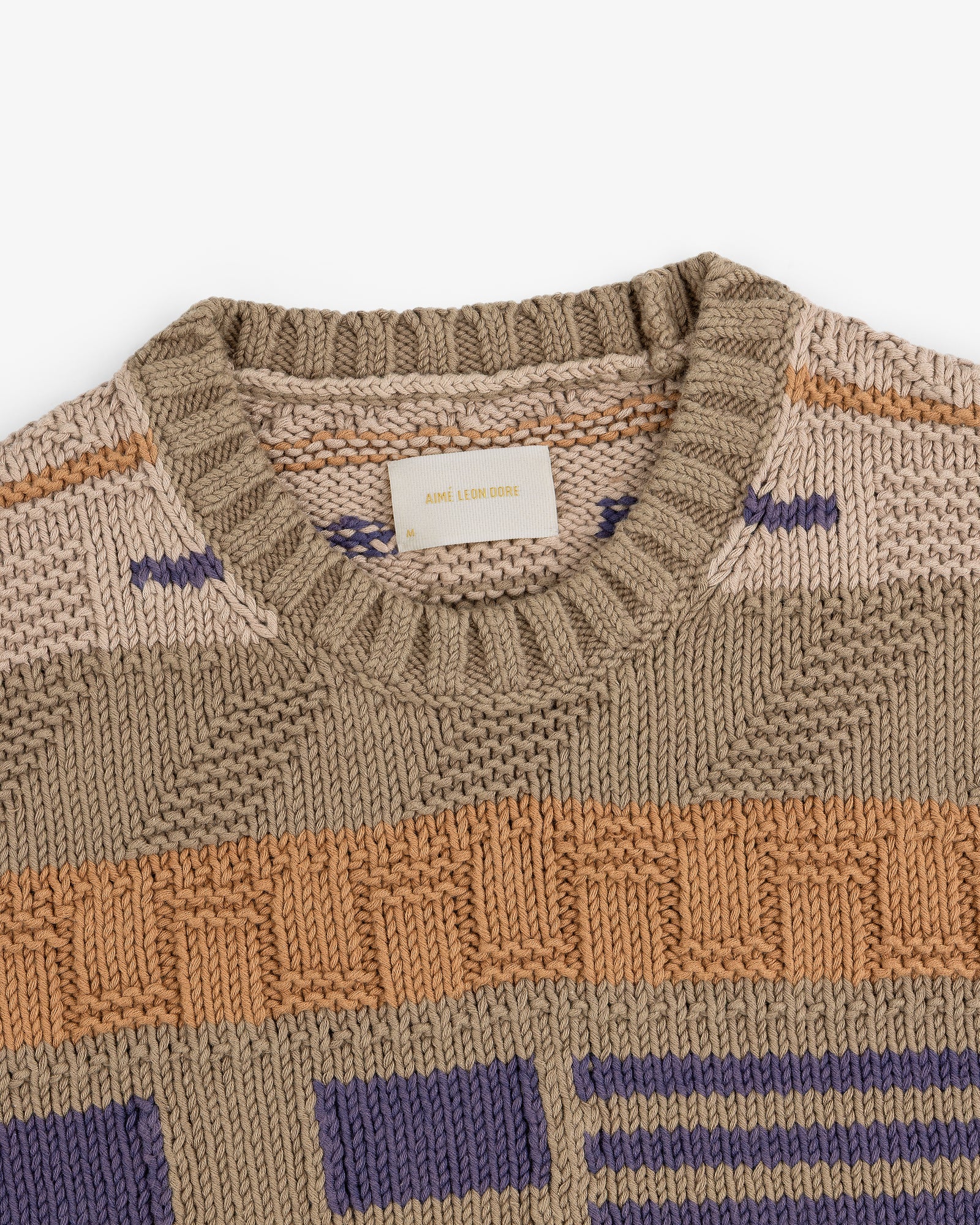 The Simaía Crewneck Sweater features beige, tan, orange, and purple geometric patterns with a ribbed crewneck collar.