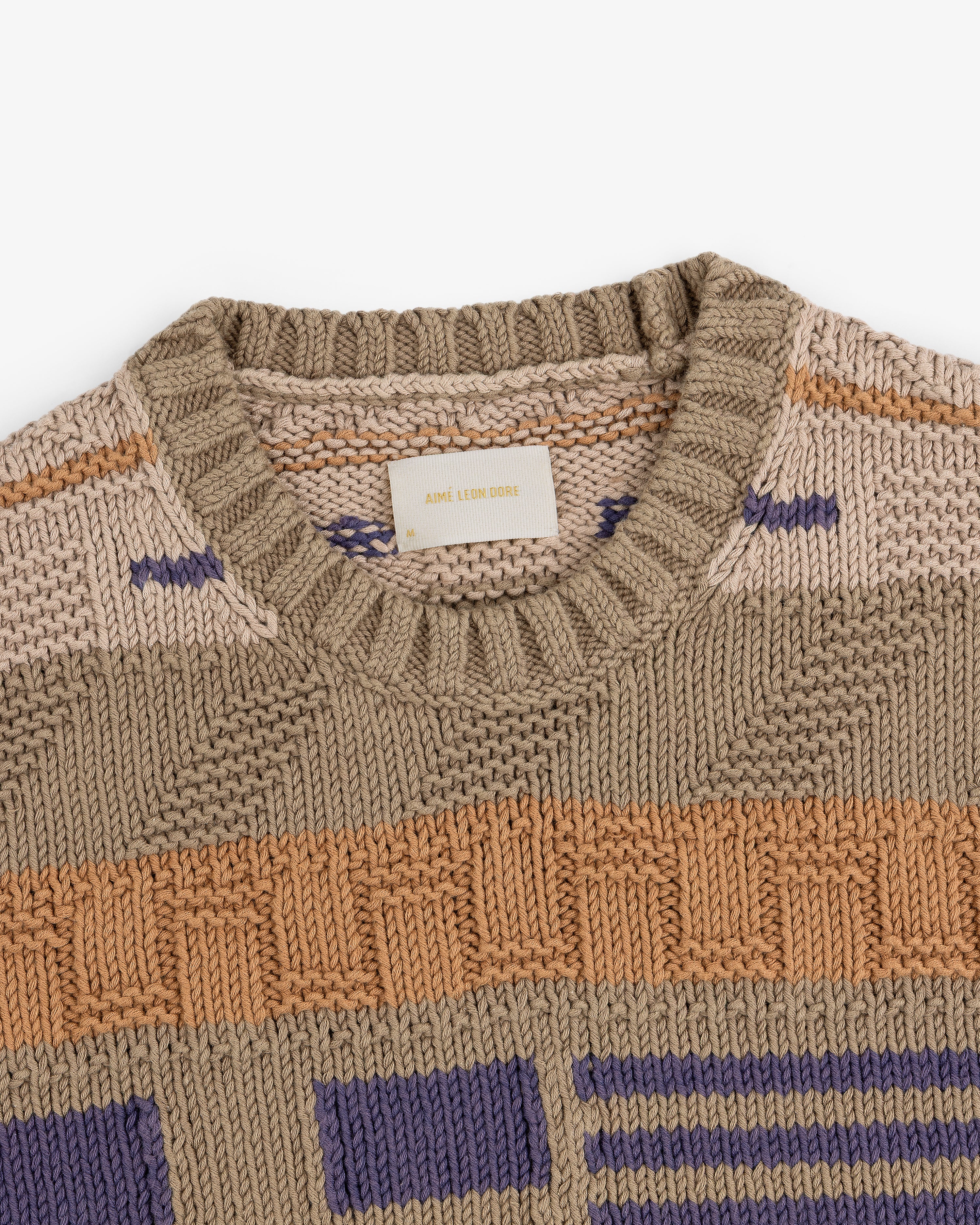 The Simaía Crewneck Sweater features beige, tan, orange, and purple geometric patterns with a ribbed crewneck collar.