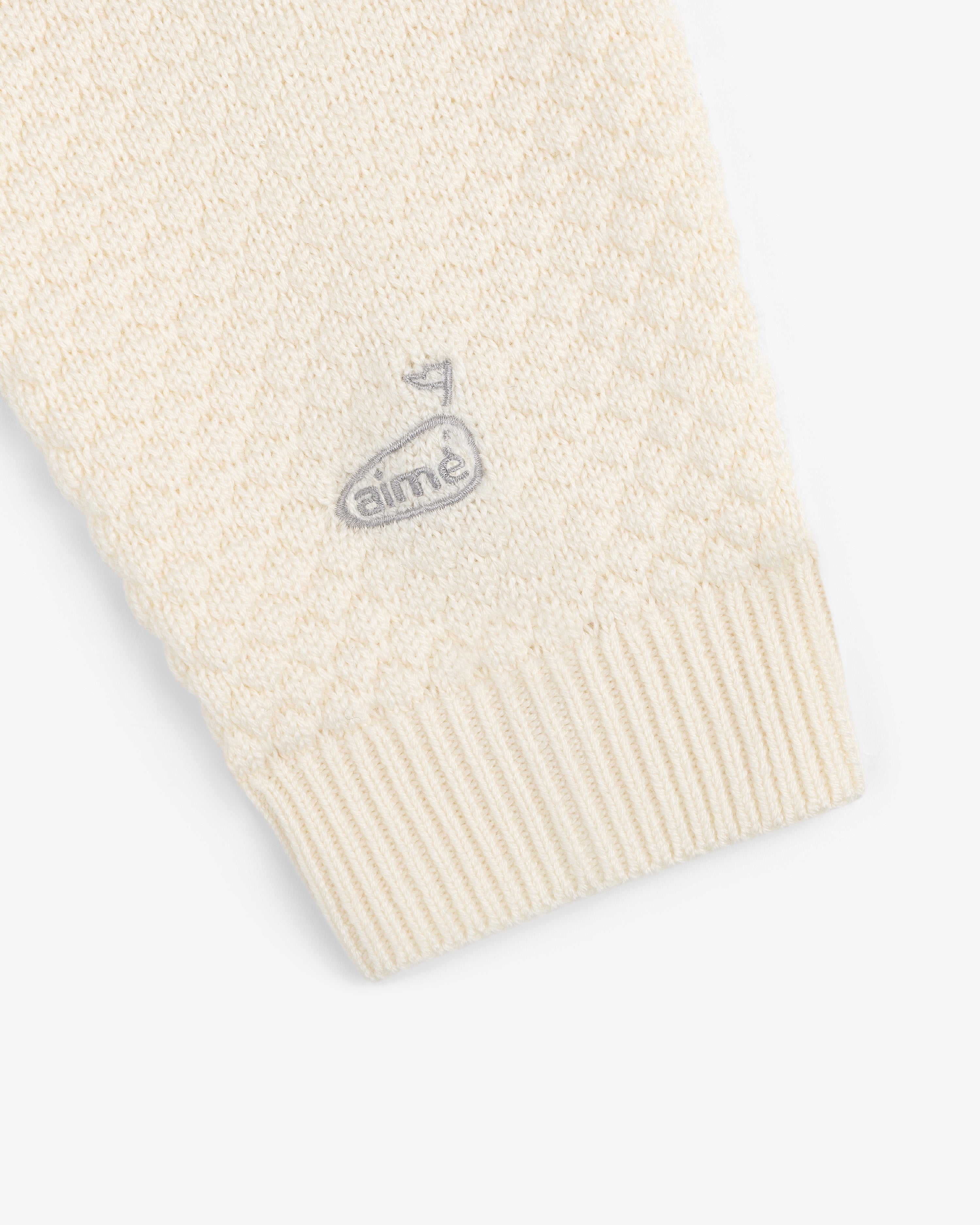 Close-up of ALD Golf Clubhouse Sweater’s cream sleeve with ribbed cuff and small embroidered golf flag logo.