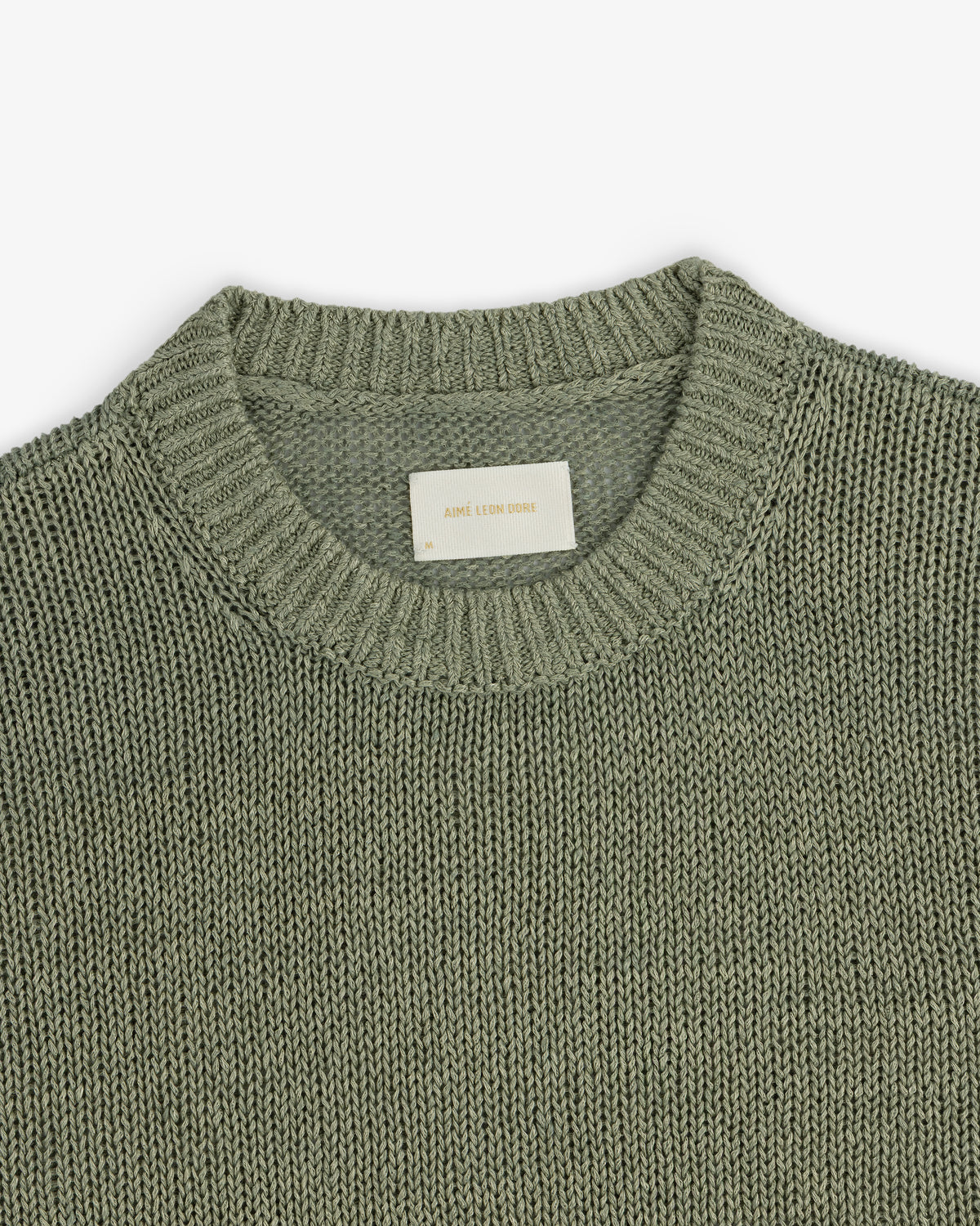 A close-up of the Linen Knit Crewneck Sweater in sage green, featuring a ribbed neckline and white inner label.