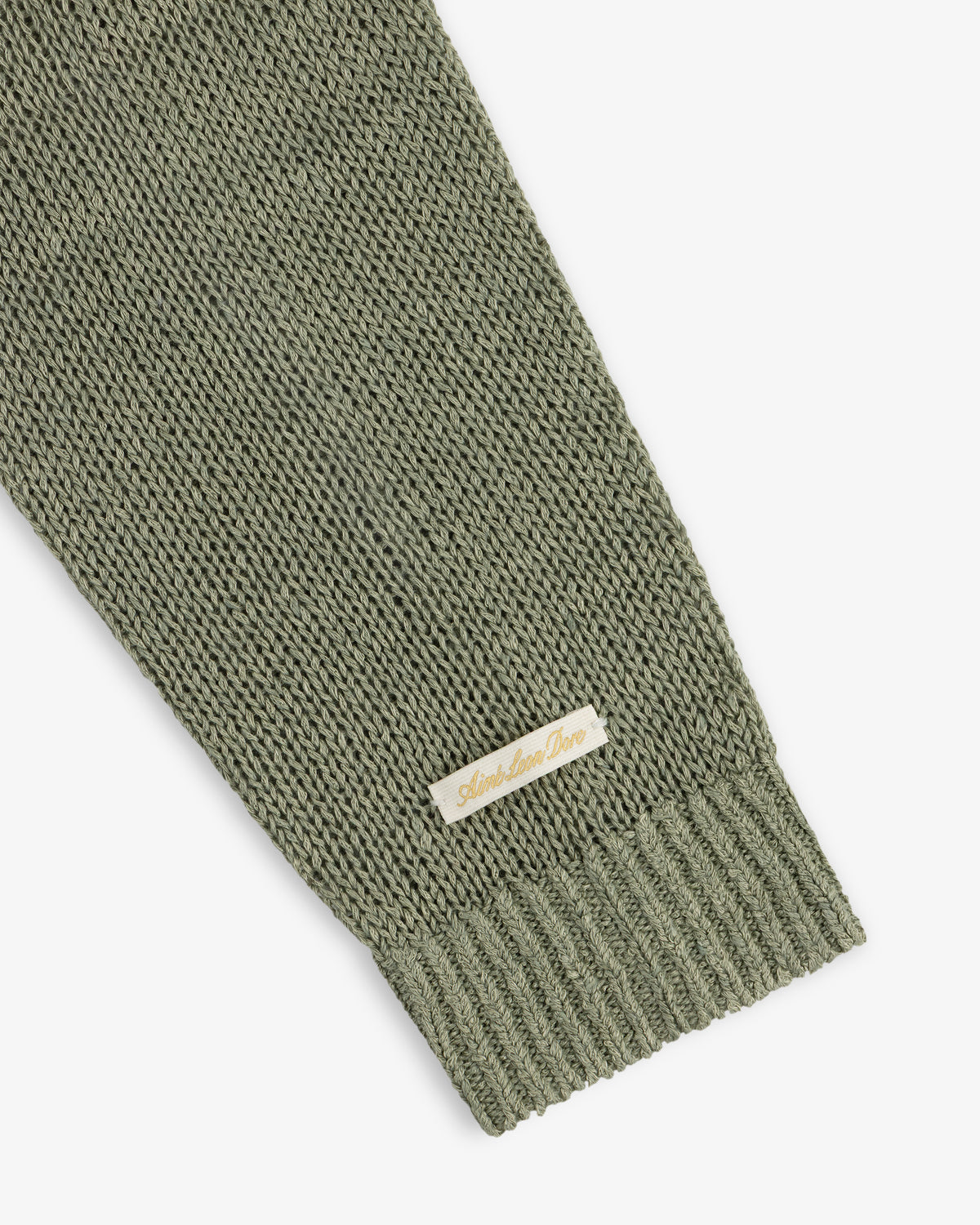 A close-up of a green Linen Knit Crewneck Sweater sleeve with a small beige Amie Amie Dare label.