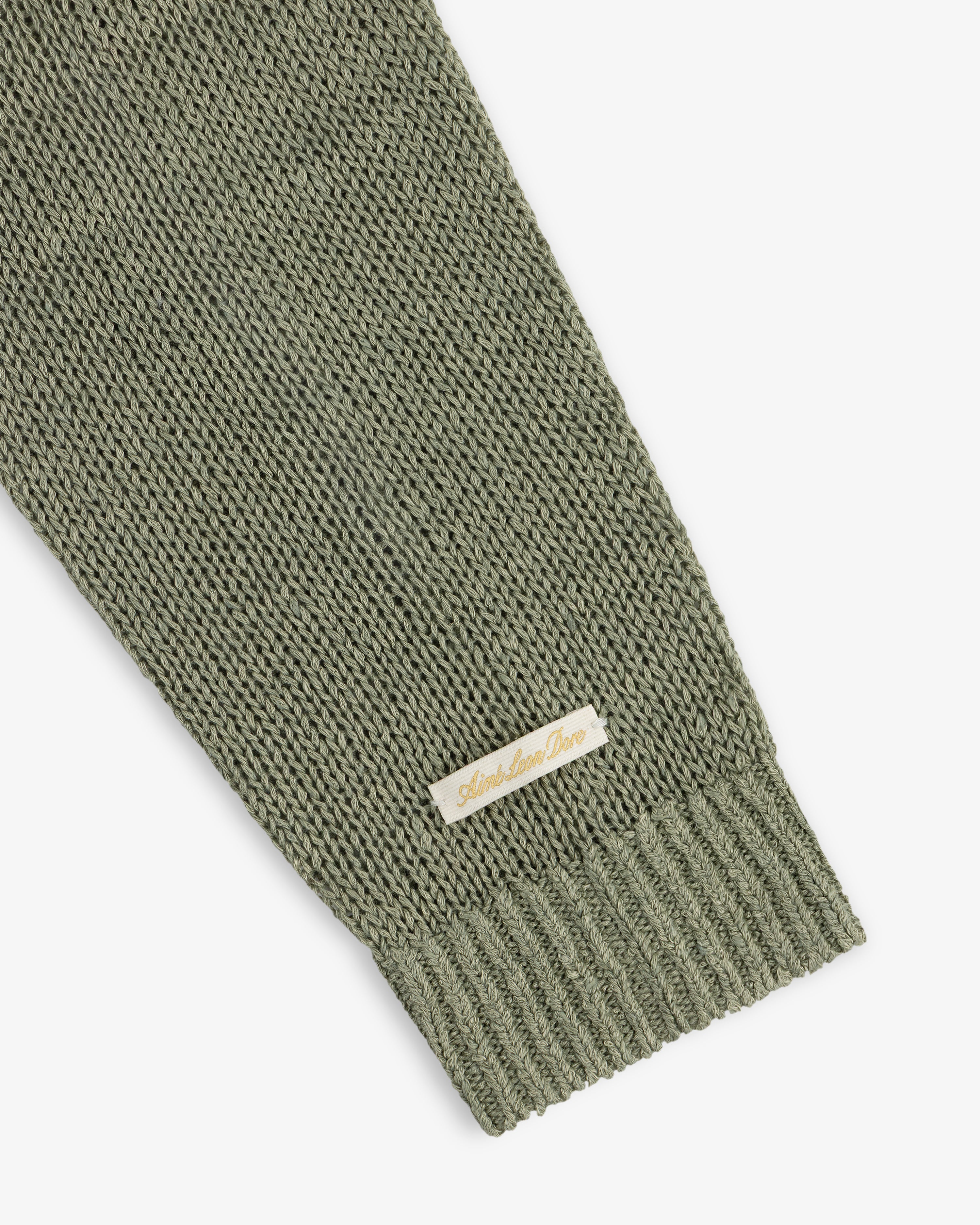 A close-up of a green Linen Knit Crewneck Sweater sleeve with a small beige Amie Amie Dare label.