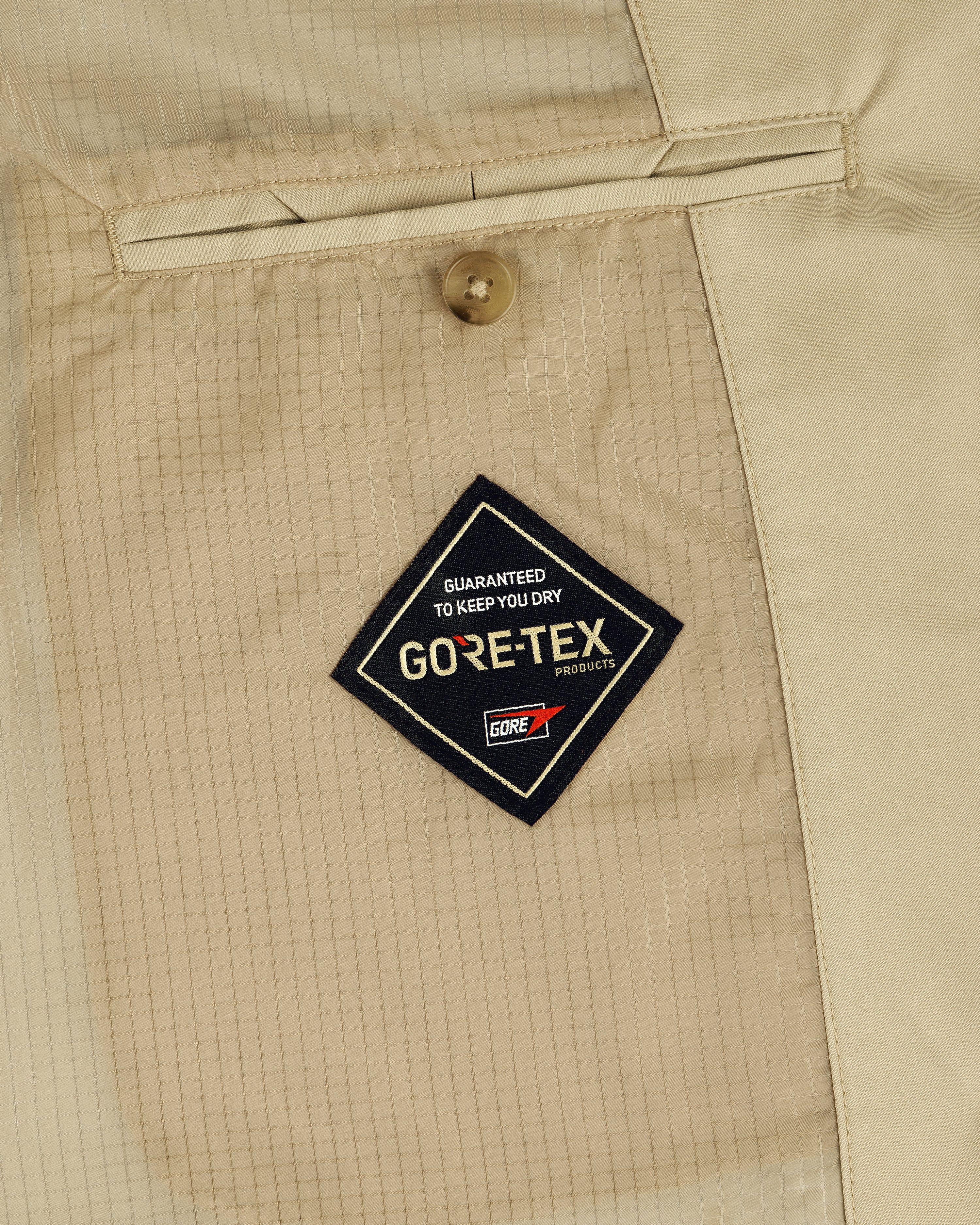 Close-up of a GORE-TEX Balmacaan Coat's beige pocket with button and logo patch reading "Guaranteed to keep you dry.