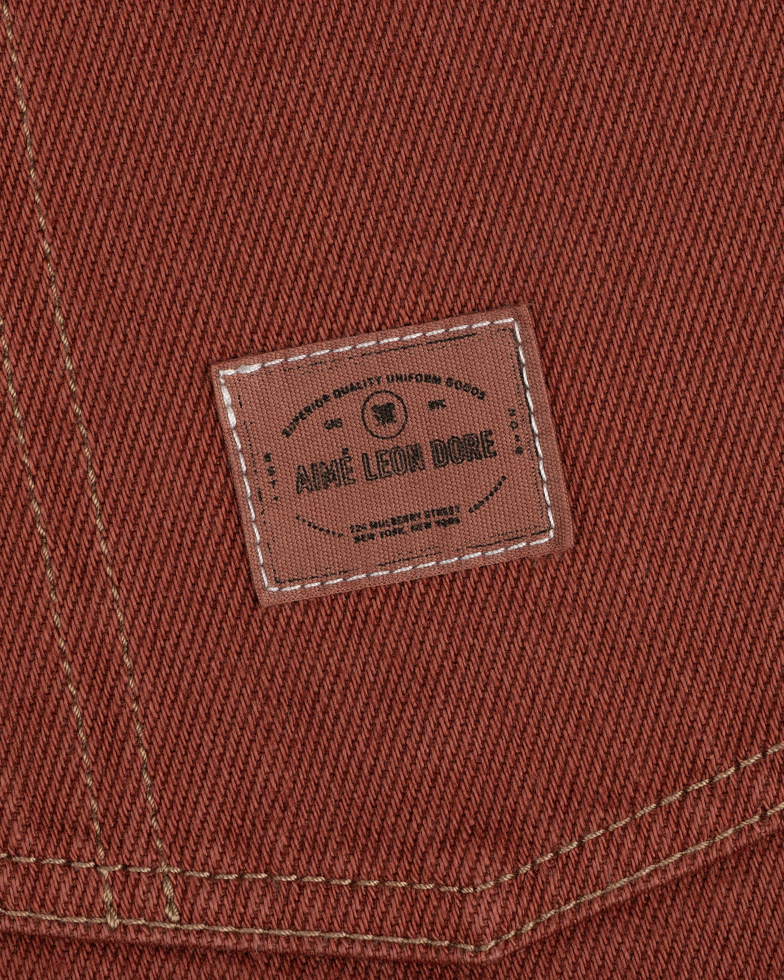 Close-up of brown fabric from the Denim Barn Jacket with a stitched Aimé Leon Dore label and white stitching.