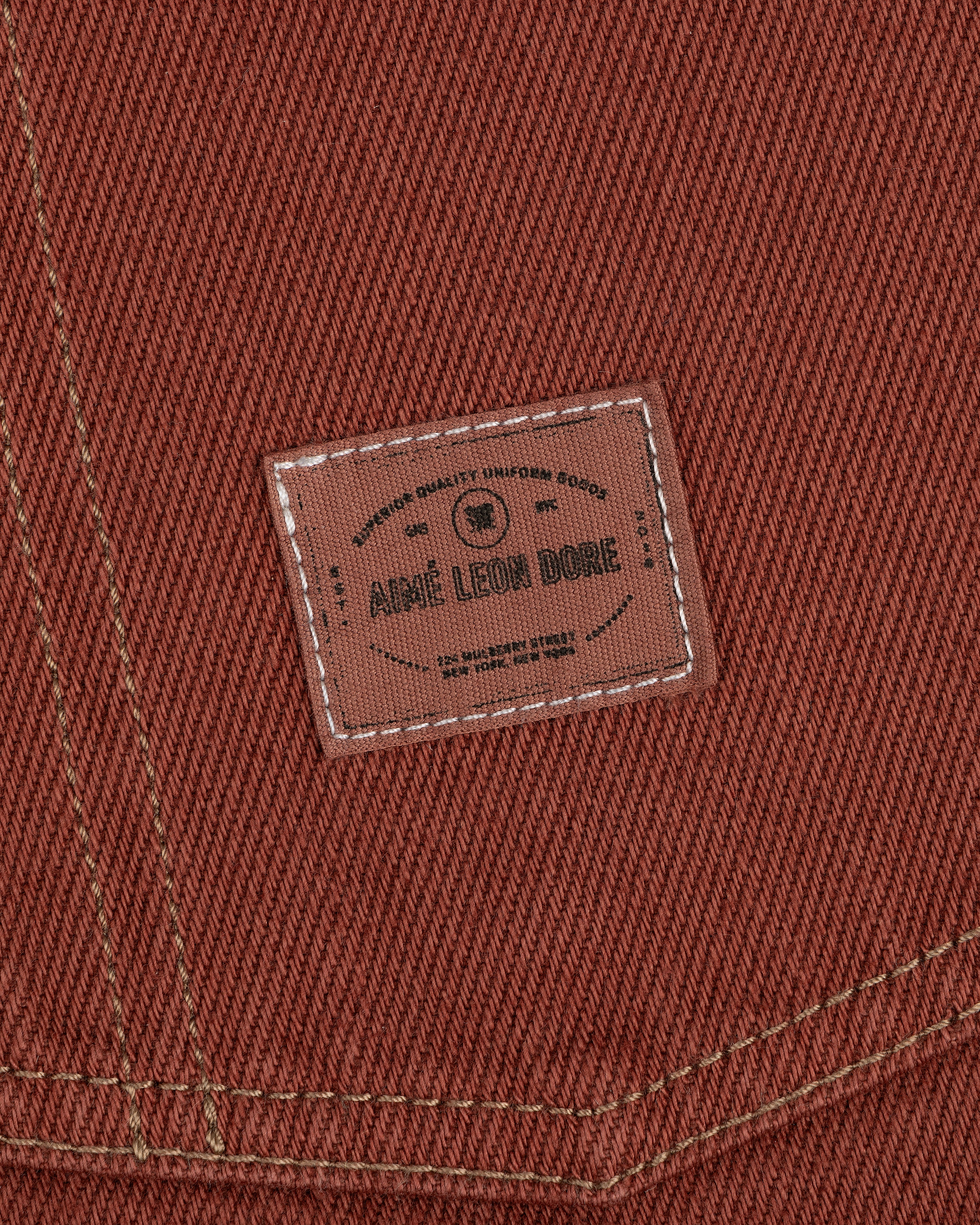 Close-up of brown fabric from the Denim Barn Jacket with a stitched Aimé Leon Dore label and white stitching.
