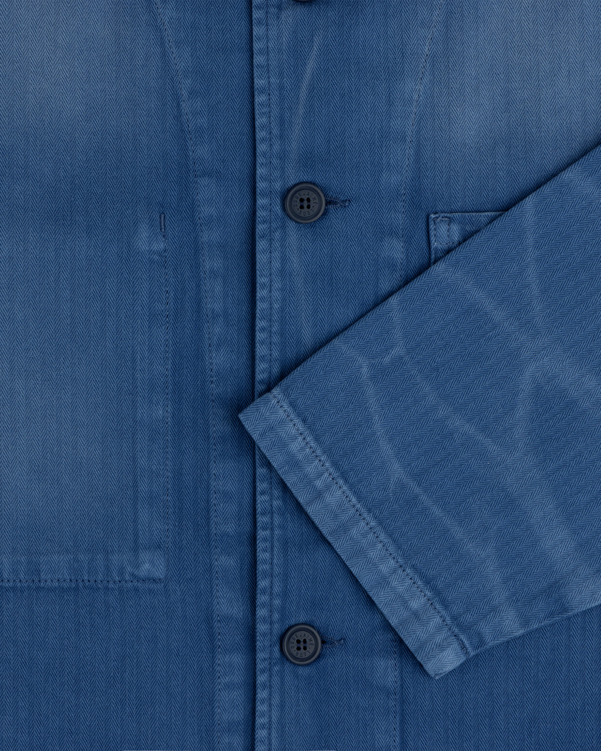 Close-up of the Sun Faded Chore Jacket, a blue button-up with a chest pocket and sleeve crossing the front.