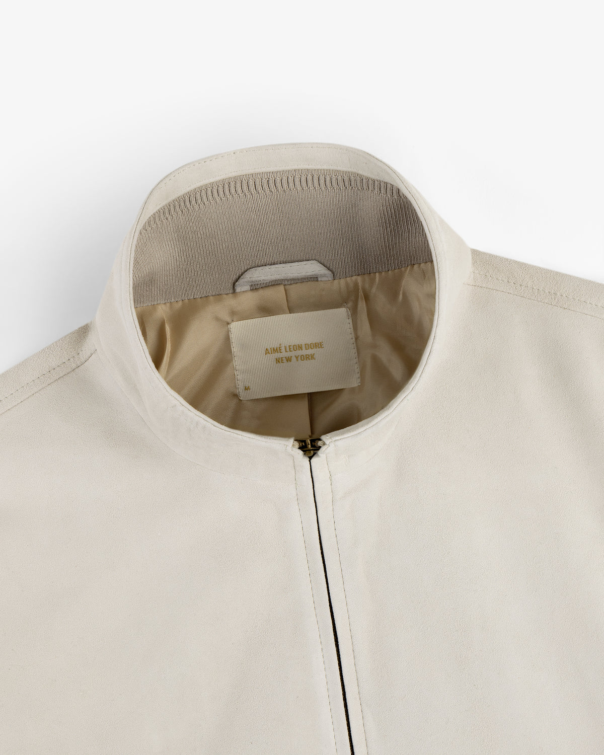 Close-up of a cream-colored Suede Track Jacket with a zipper and a Jane Léon Dioré New York label inside the collar.