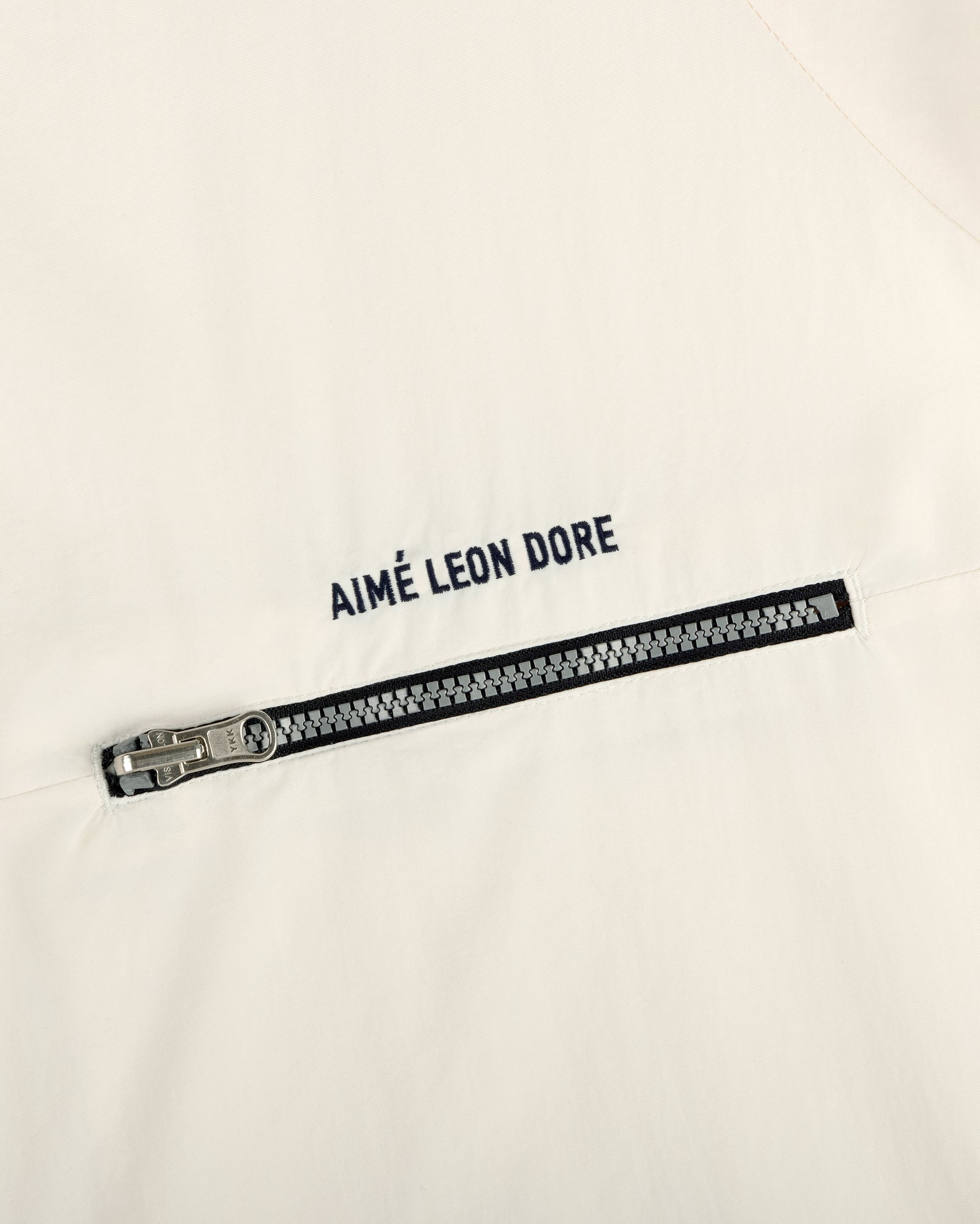 Close-up of an off-white Nylon Packable Windbreaker with a silver zipper and AIMÉ LEON DORE text above it.