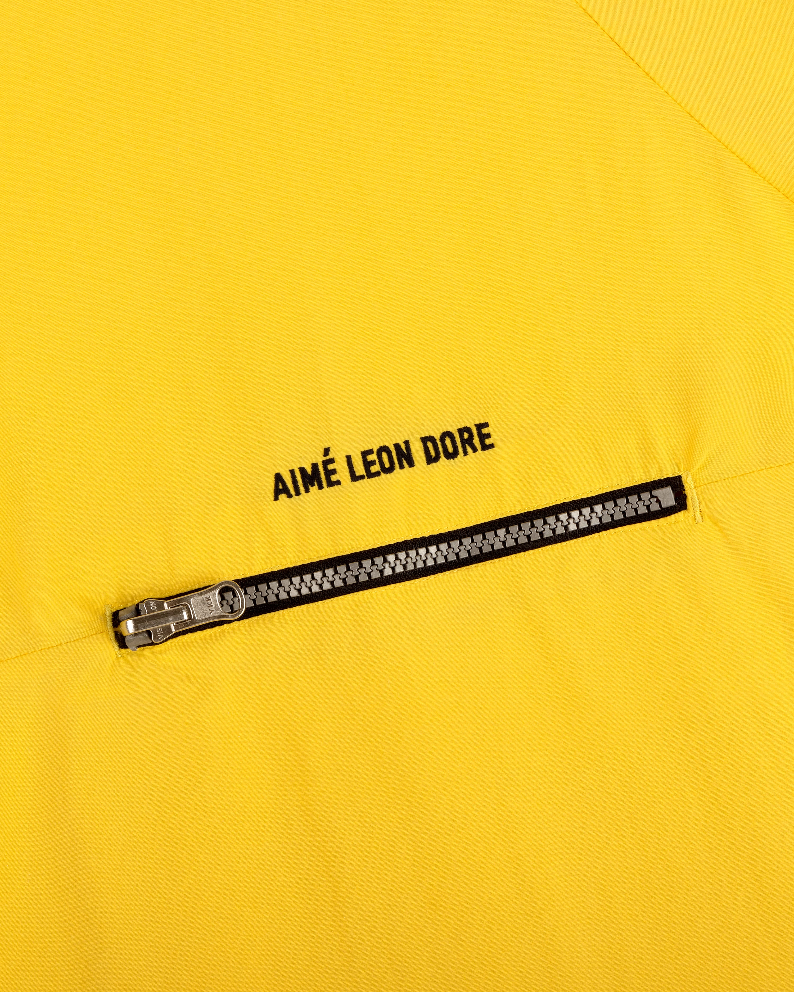 Close-up of the Nylon Packable Windbreaker in yellow, featuring a diagonal zipper and AIMÉ LEON DORE text above.