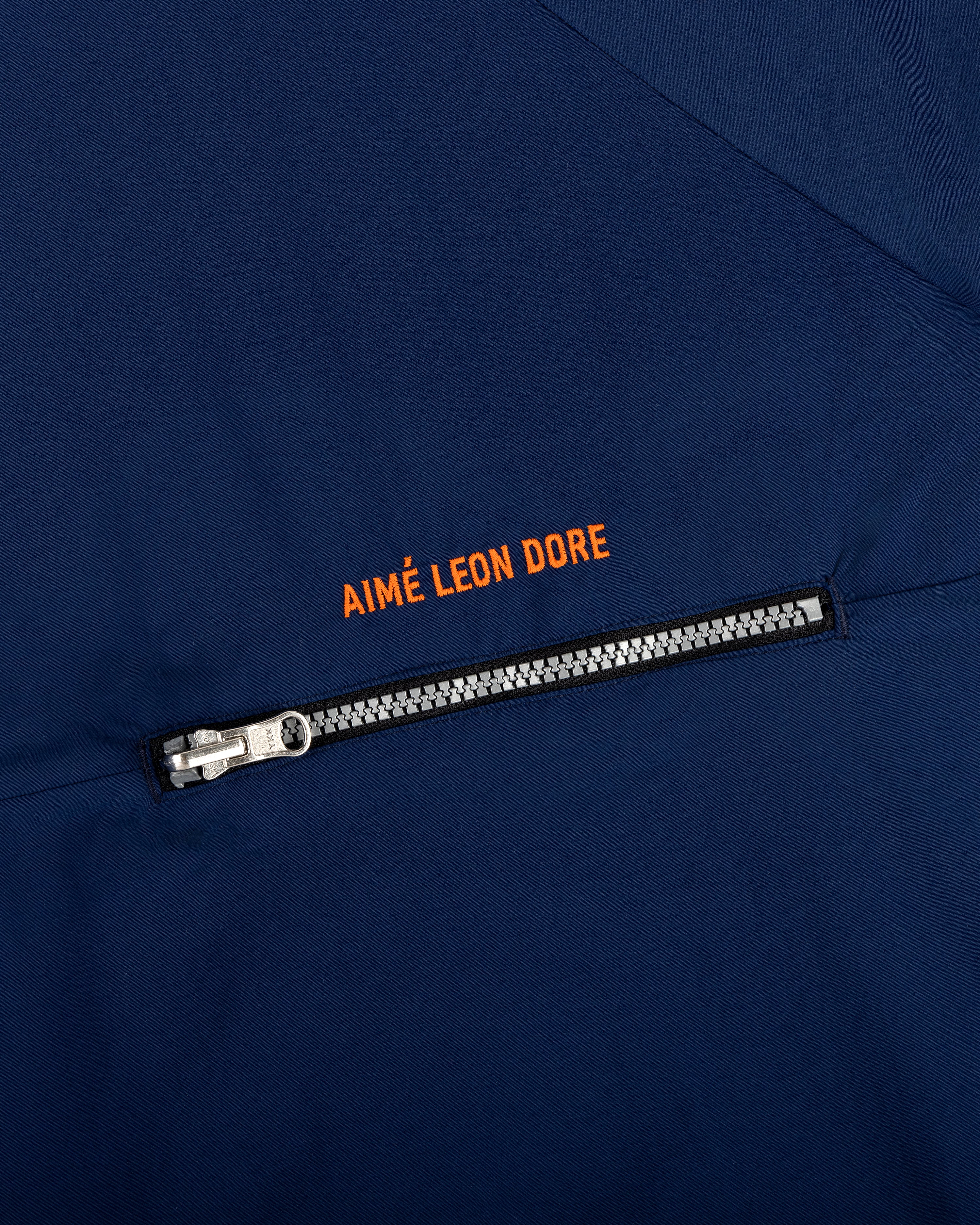 Close-up of the Nylon Packable Windbreaker in navy with a silver zipper and orange AIMÉ LEON DORE text above it.