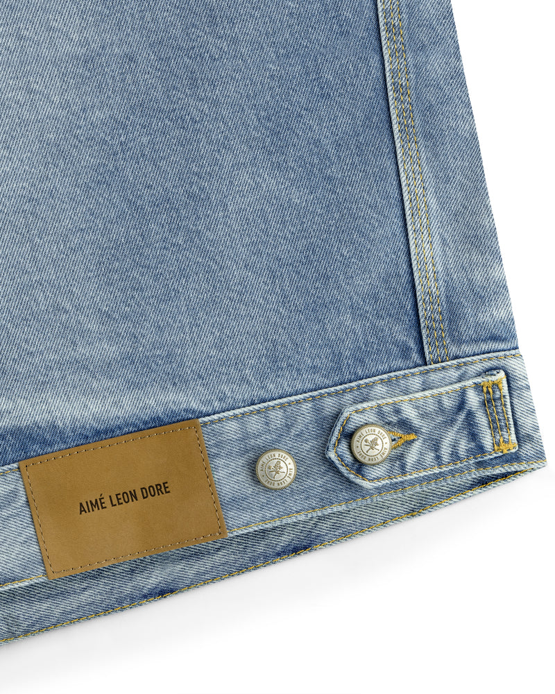 Close-up of light blue Denim Trucker Jacket featuring buttons and a tan leather patch labeled AIMÉ LEON DORE.