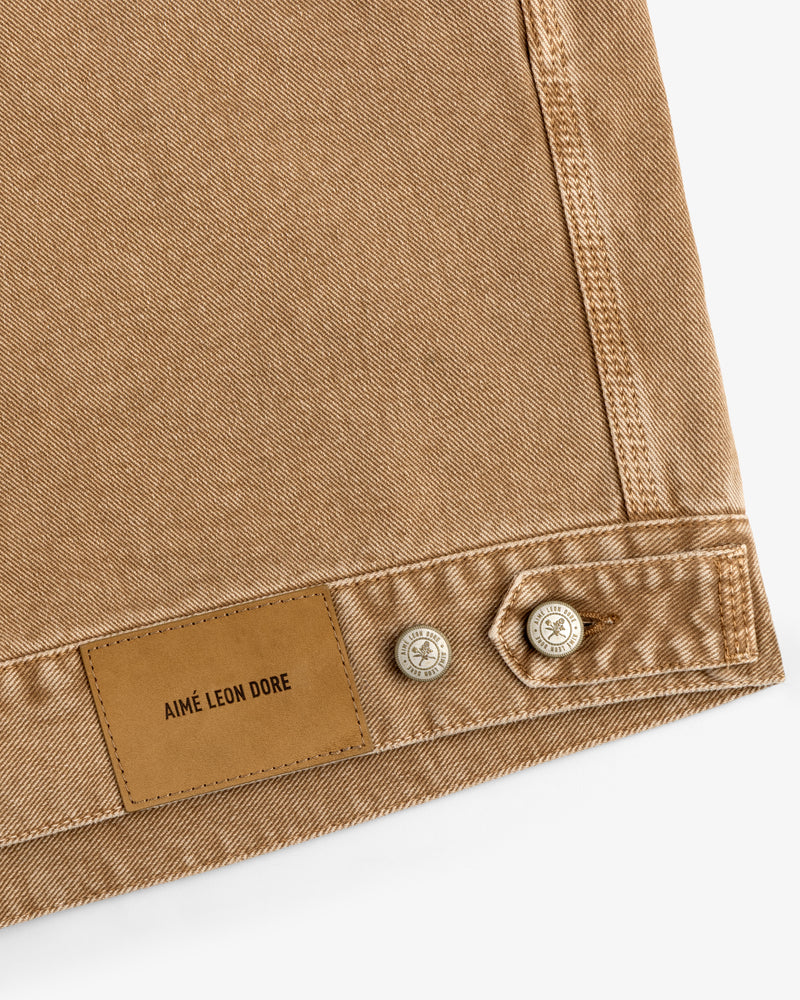 Close-up of tan Denim Trucker Jacket fabric with leather Aimé Leon Dore patch and two metal buttons on white background.
