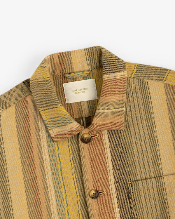 The Blanket Striped Chore Jacket features earth-tone stripes, a collar, brown buttons, and a front chest pocket.