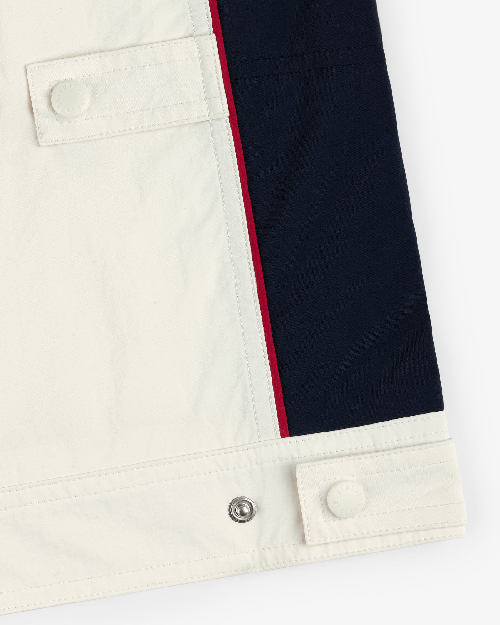 Close-up of the Nylon Moto Jacket hem with white and navy fabric, red stripe, and button tab details.