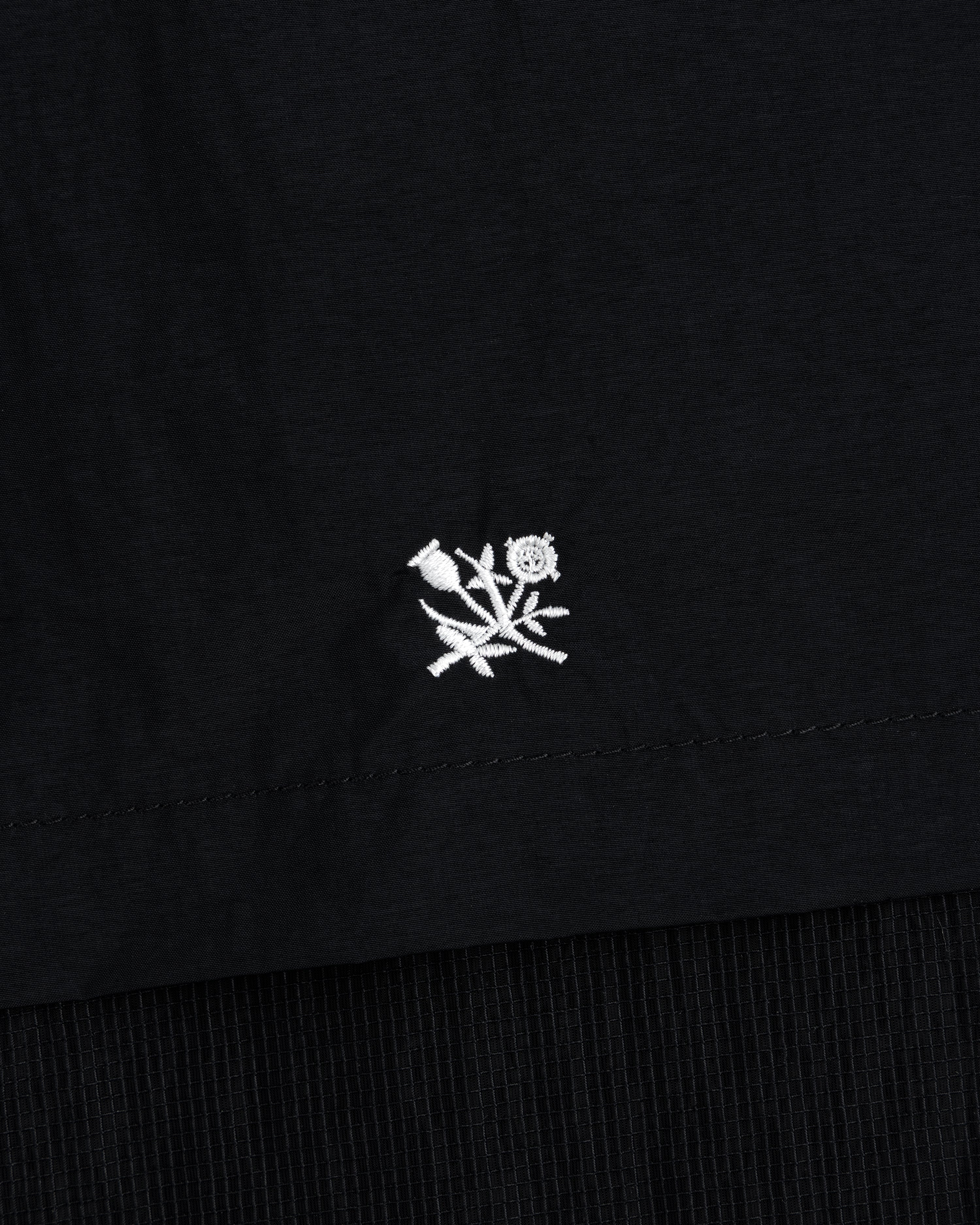 Small white embroidered flower near the hem of a black Nylon Track Jacket.