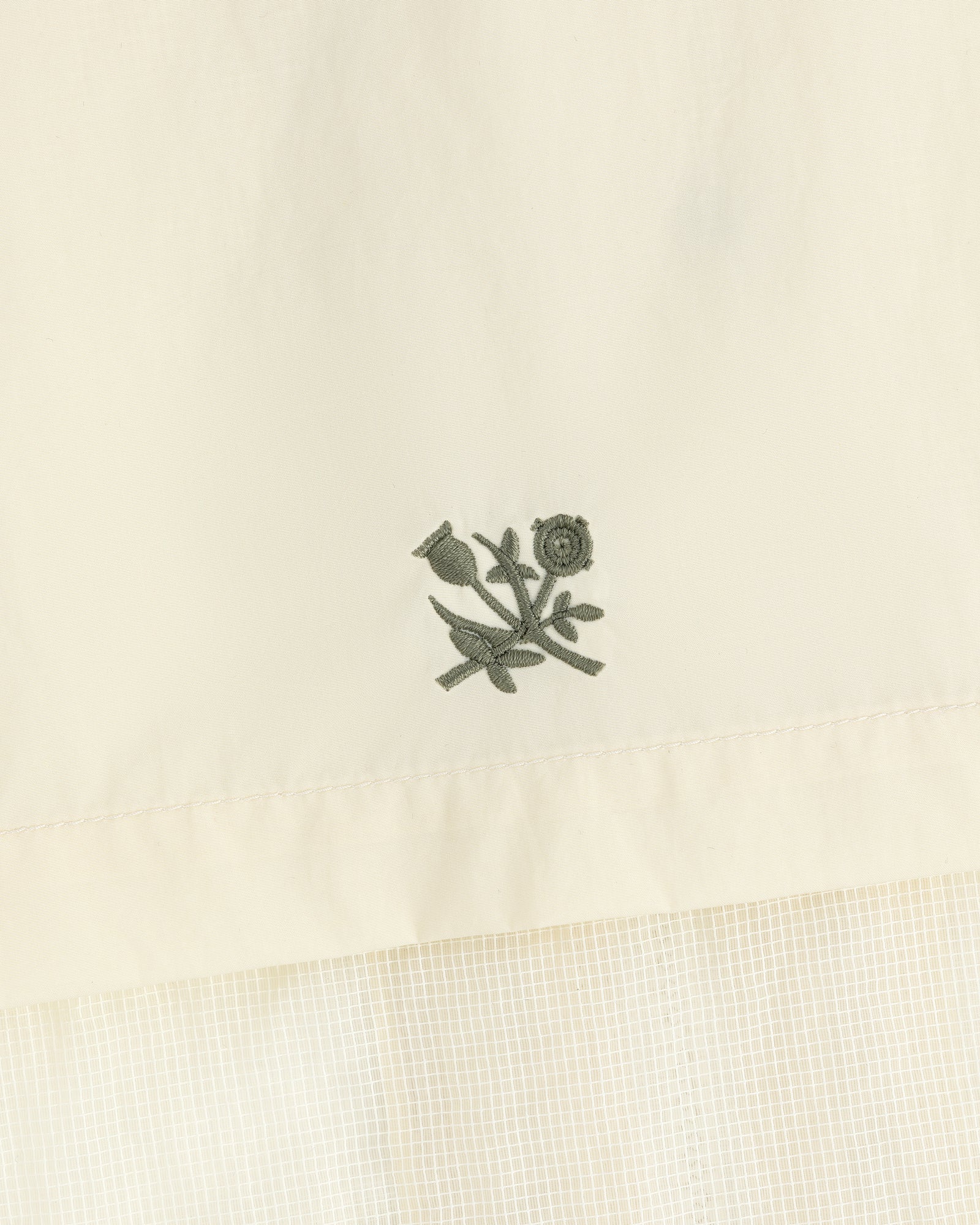 Embroidered green flower and stem motif on cream nylon track jacket with a subtle grid pattern below.