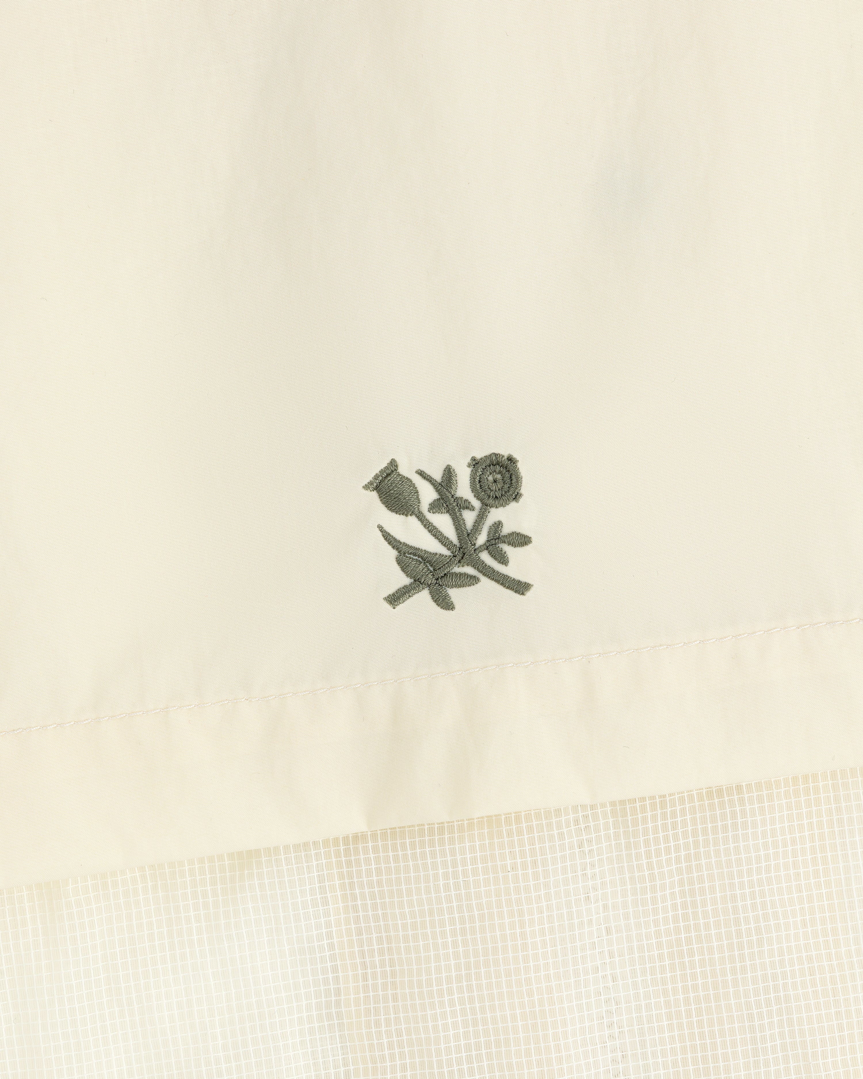 Embroidered green flower and stem motif on cream nylon track jacket with a subtle grid pattern below.
