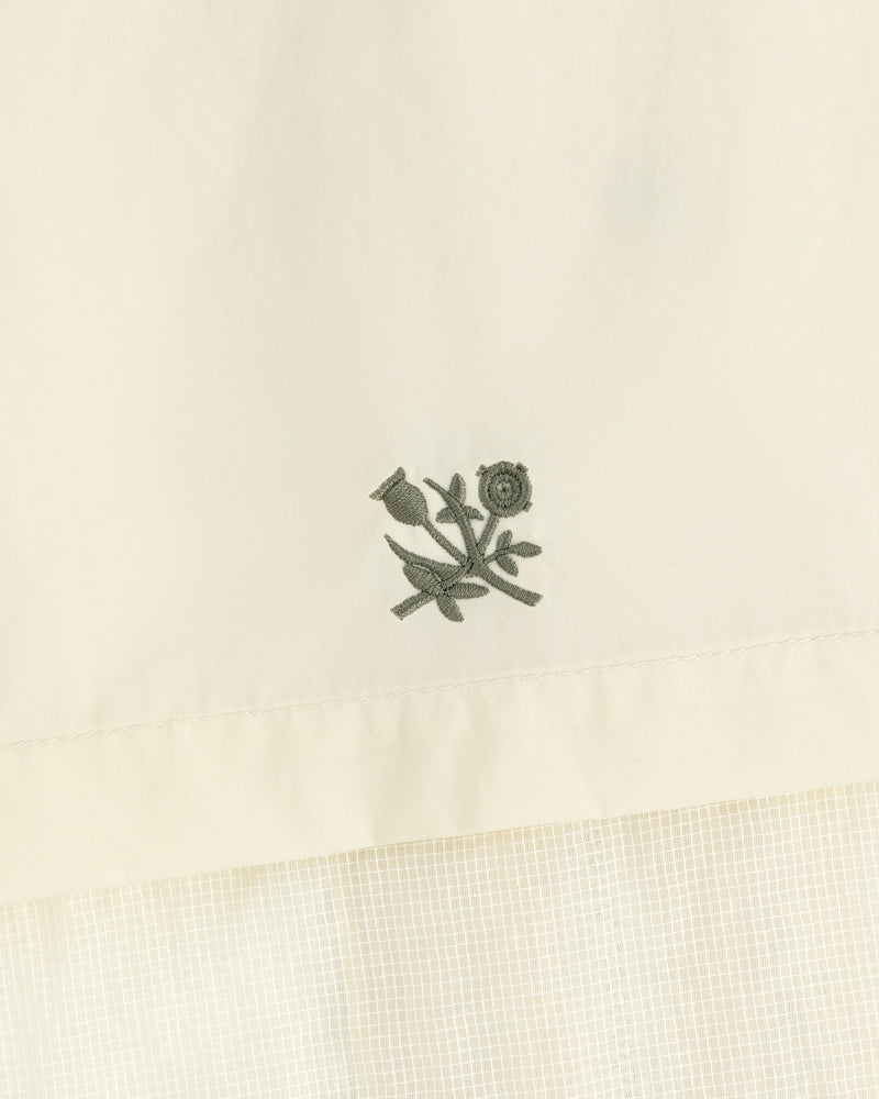 Embroidered green flower and stem motif on cream nylon track jacket with a subtle grid pattern below.