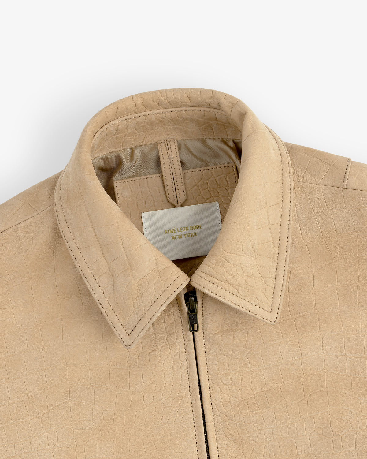 Close-up of a beige Croc-Embossed Nubuck Bomber Jacket with collar and zipper, label reads AIME LEON DORE NEW YORK.