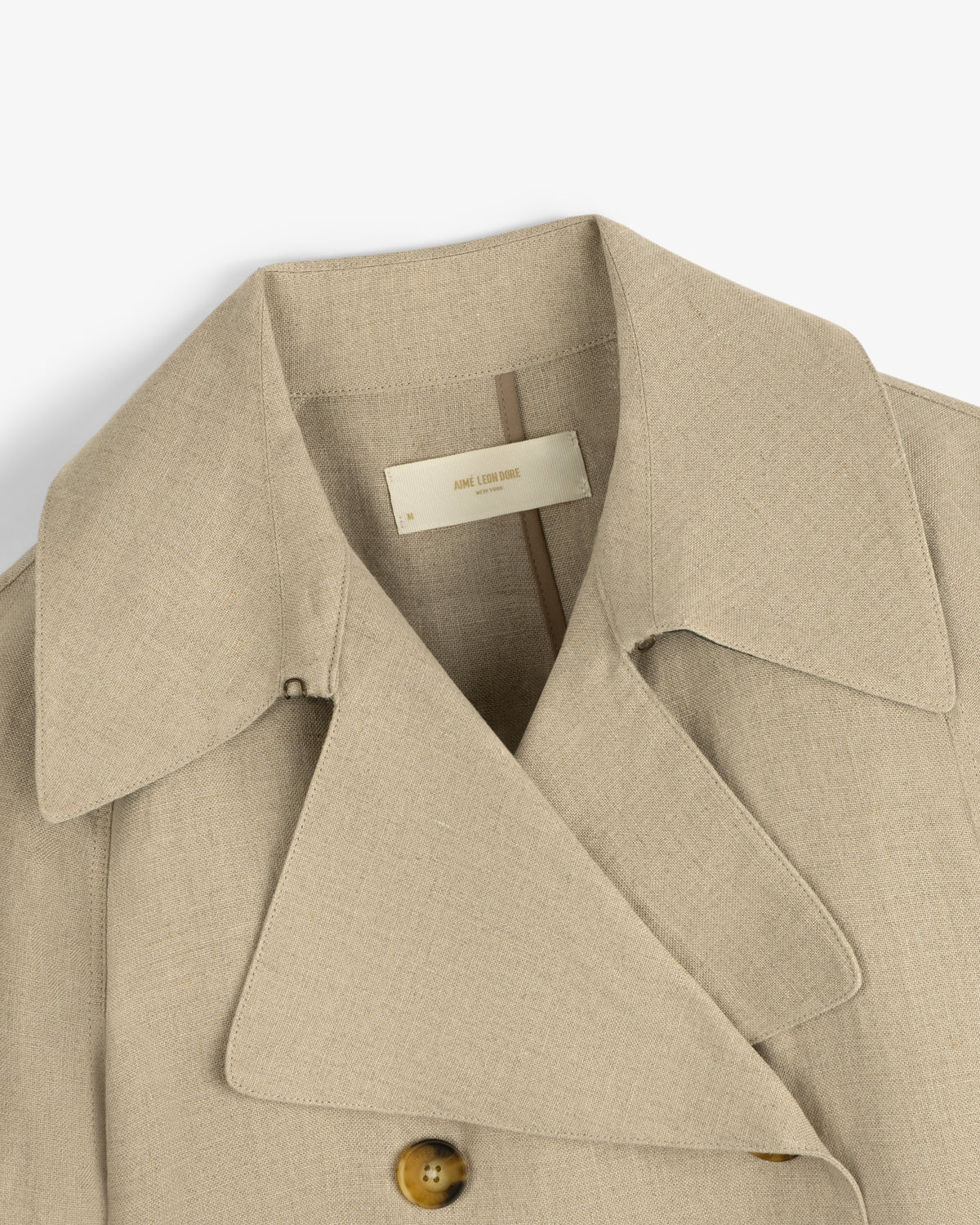 Close-up of the Linen Trench Coat in beige with wide lapels, brown button, and visible inside label.