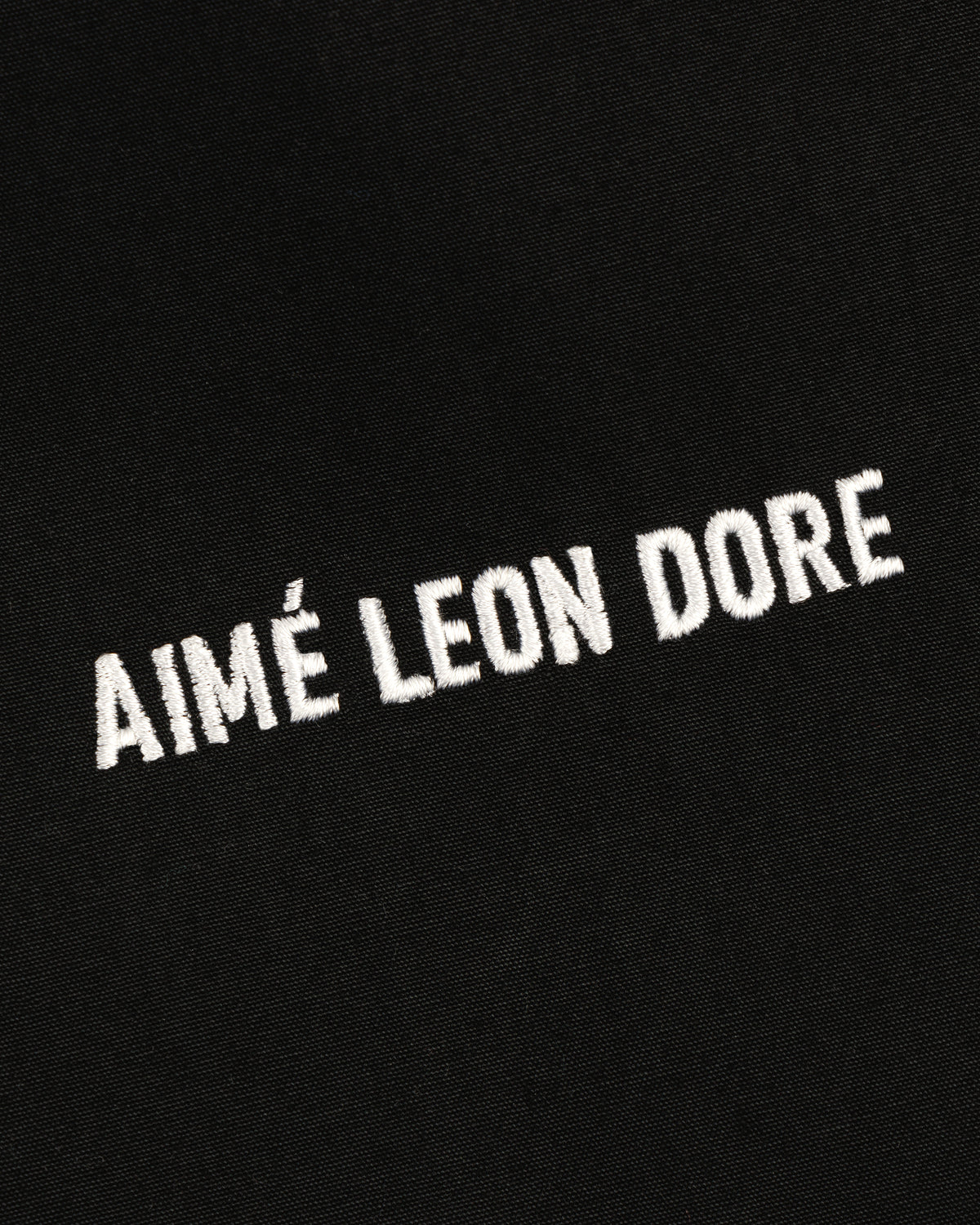 White Micro Logo embroidery on black fabric of the Lightweight Jacket, shown at a slight diagonal angle.