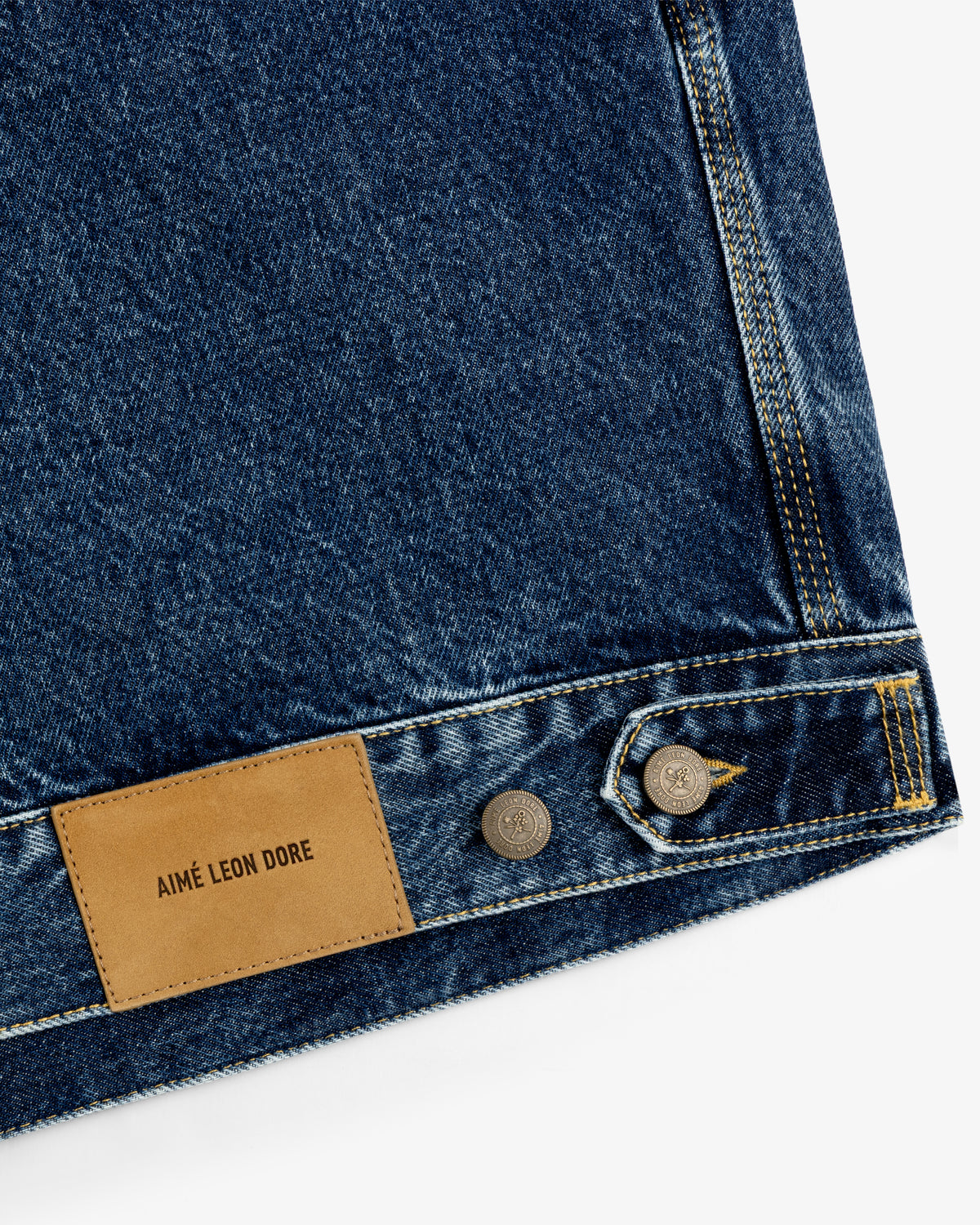 Close-up of blue Denim Trucker Jacket with AIMÉ LEON DORE leather patch and two metal buttons.