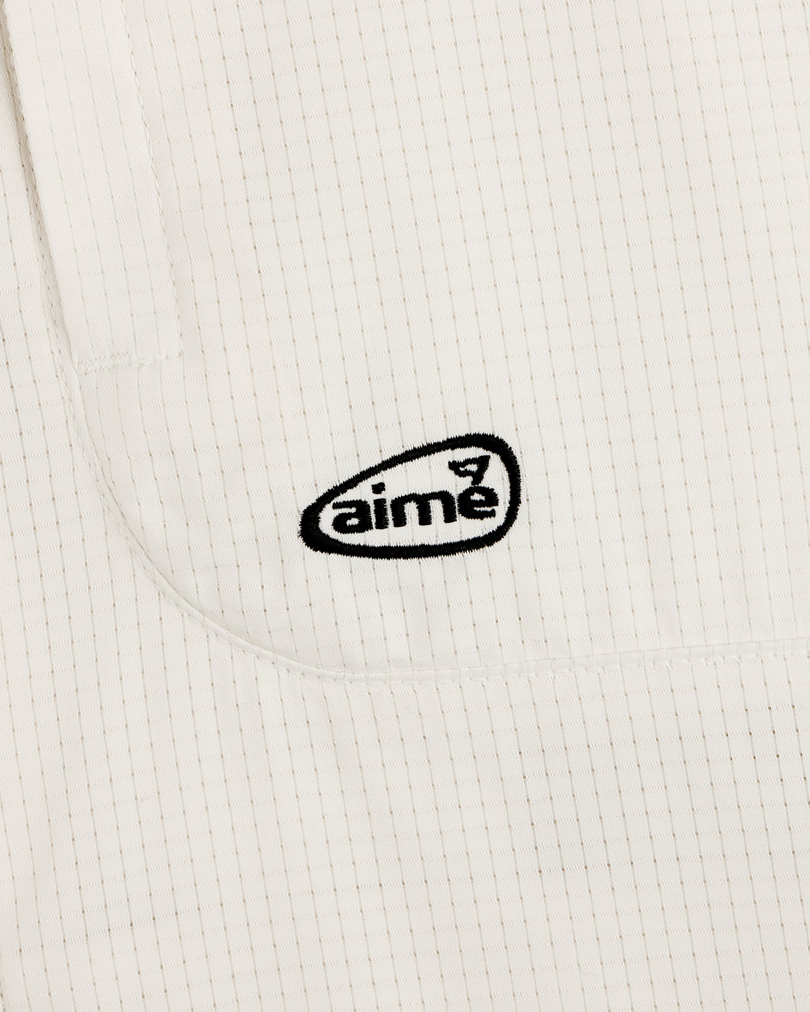 Close-up of the ALD Golf Lightweight Tips Windbreaker: white fabric with thin vertical stripes and black aimé logo embroidery.