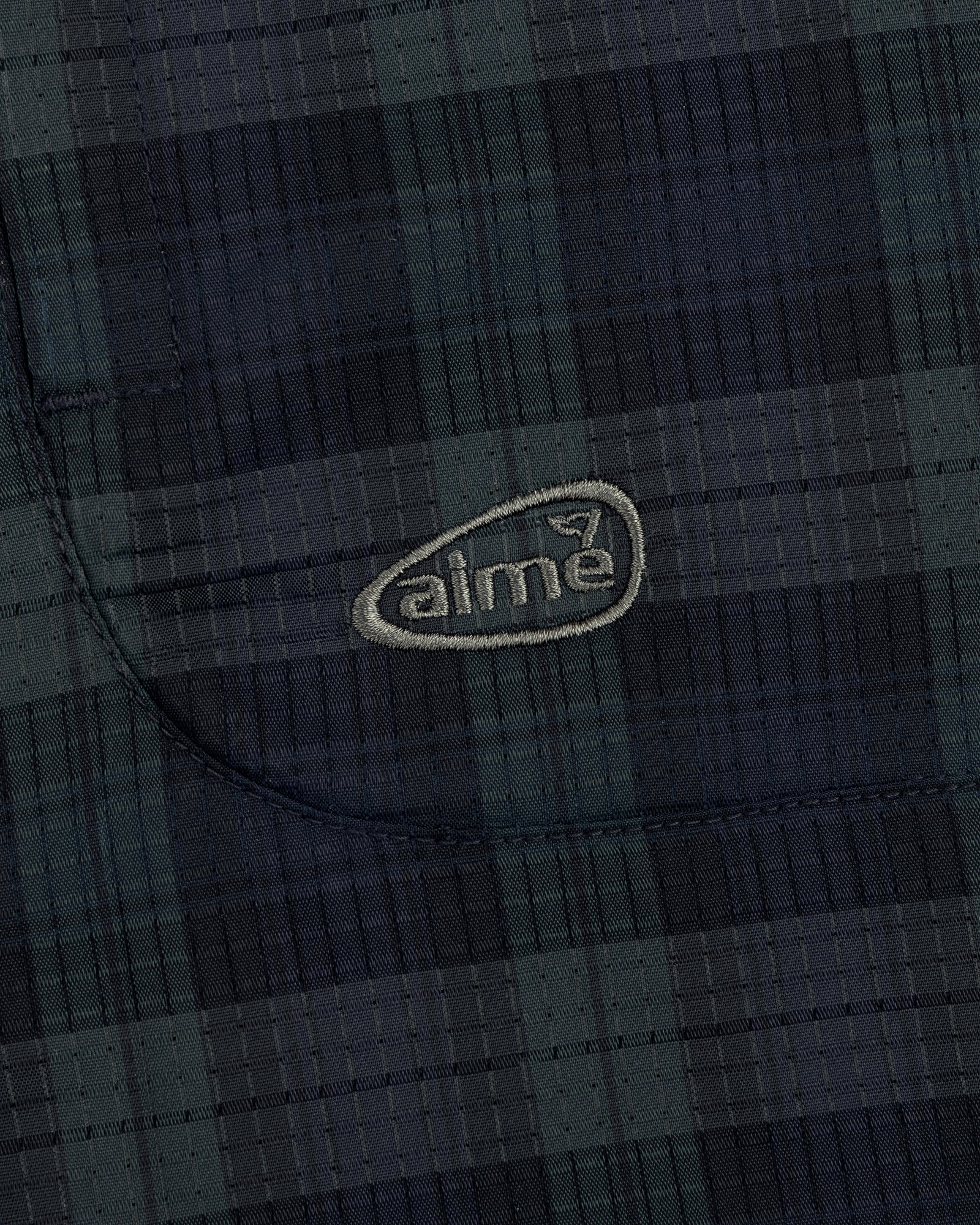 Close-up of a dark green and navy plaid ALD Golf Lightweight Tips Windbreaker with "aime" embroidered in light thread.